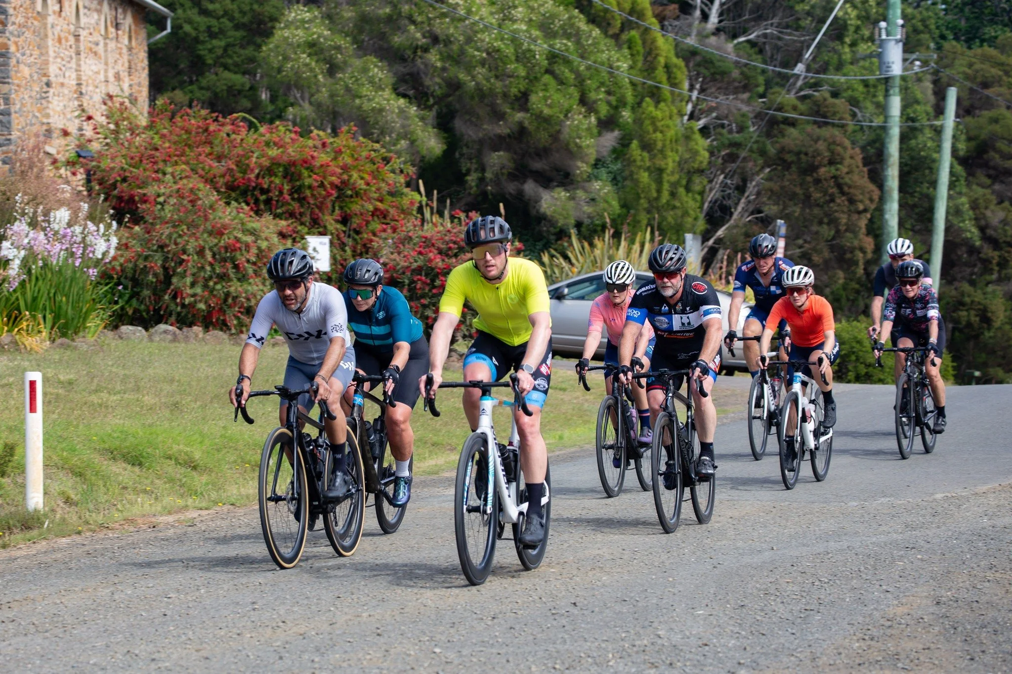 Group of eight cyclists riding on a rural road surrounded by greenery and shrubs.