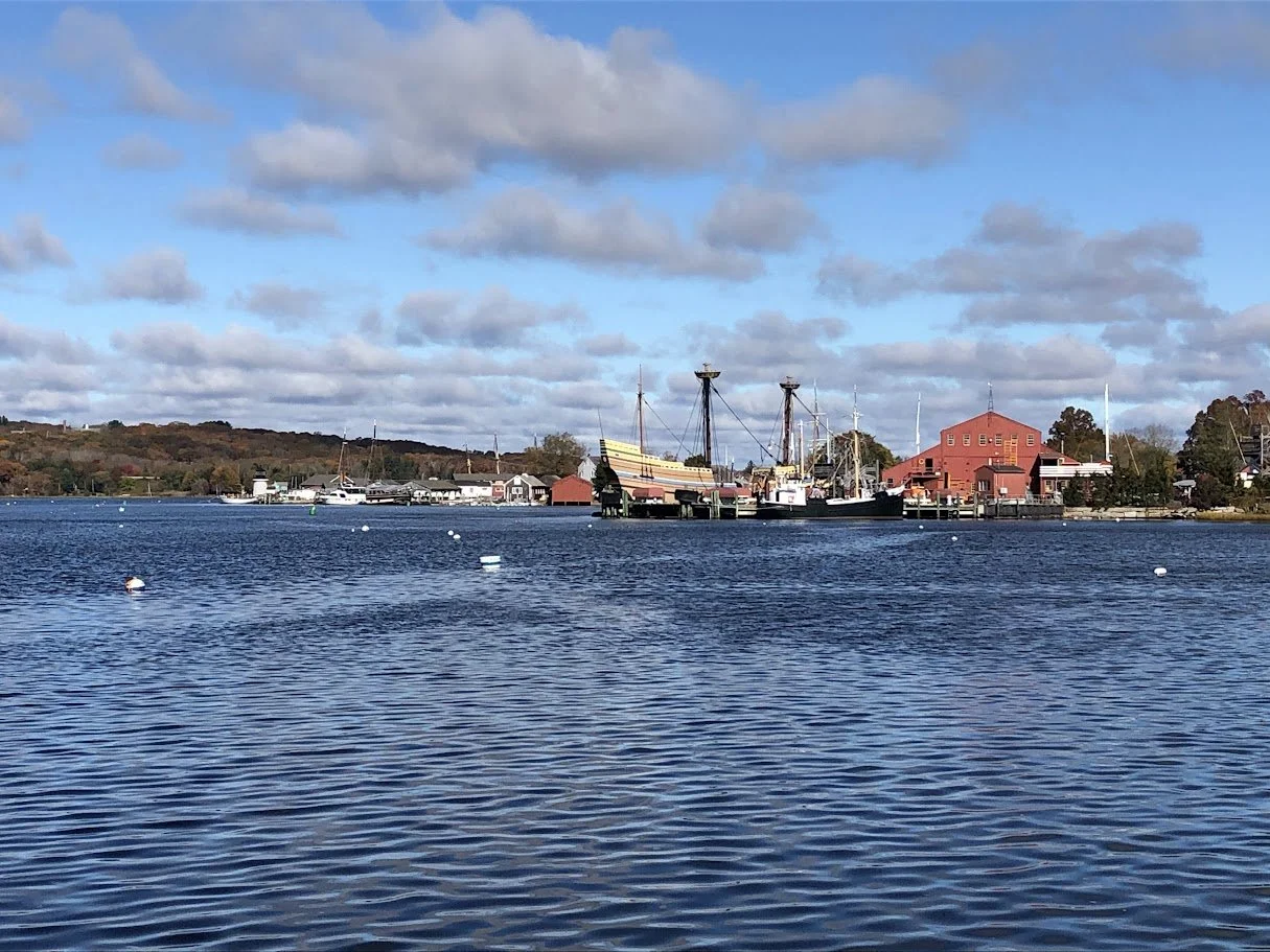 A harbor with boats, including a large ship resembling a pirate or historical vessel, docked near red and white buildings. The water is calm and reflects the sky, with a partly cloudy sky overhead.