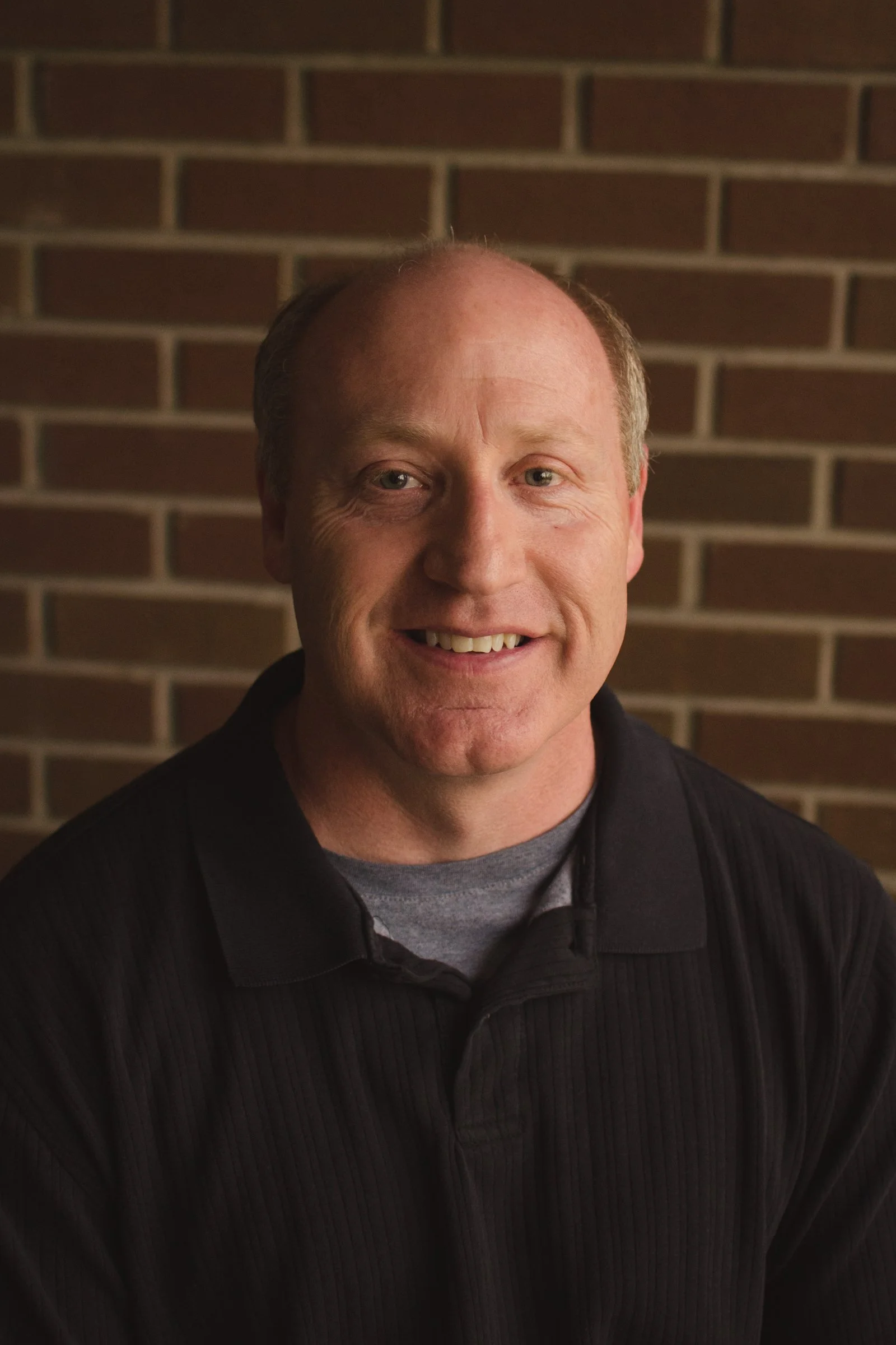 A middle-aged man with a friendly smile, light skin, short light-colored hair, wearing a black jacket and a gray shirt, standing in front of a brick wall.