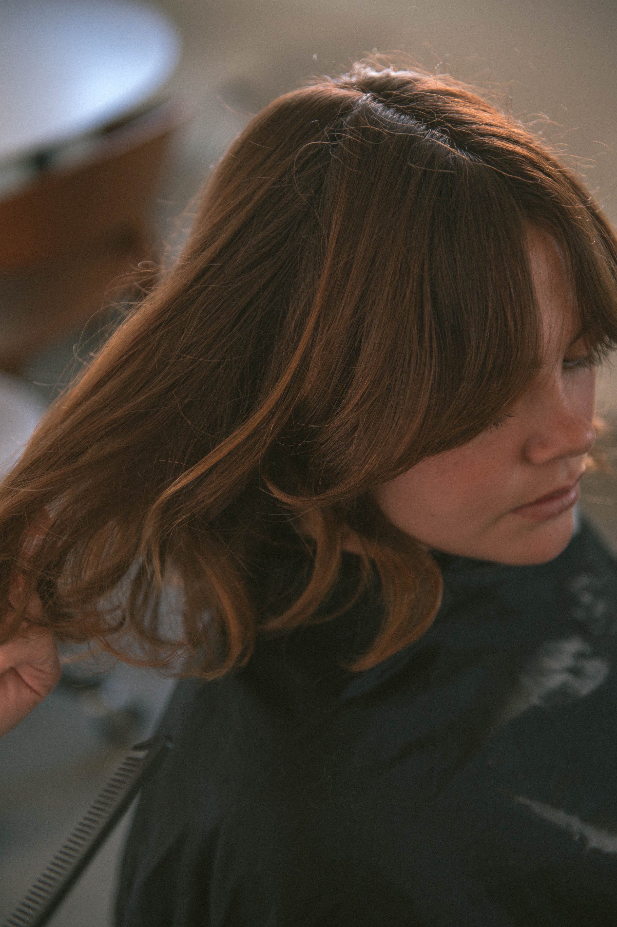 A woman with brown, wavy hair and fair skin sitting at a salon, getting her hair styled.