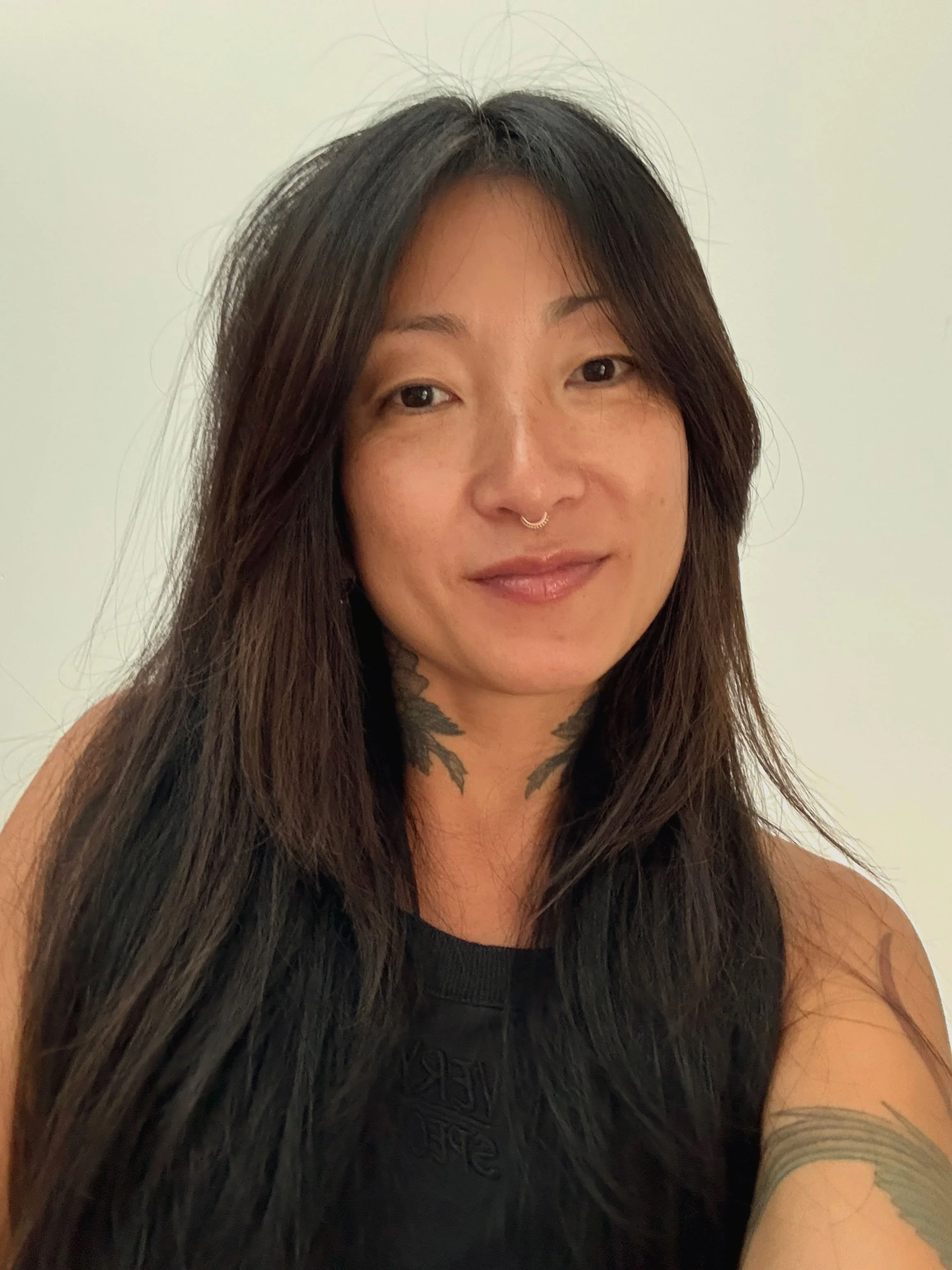 A woman with long dark hair, wearing a black sleeveless shirt, with tattoos on her neck and arm, smiling slightly against a plain white background.
