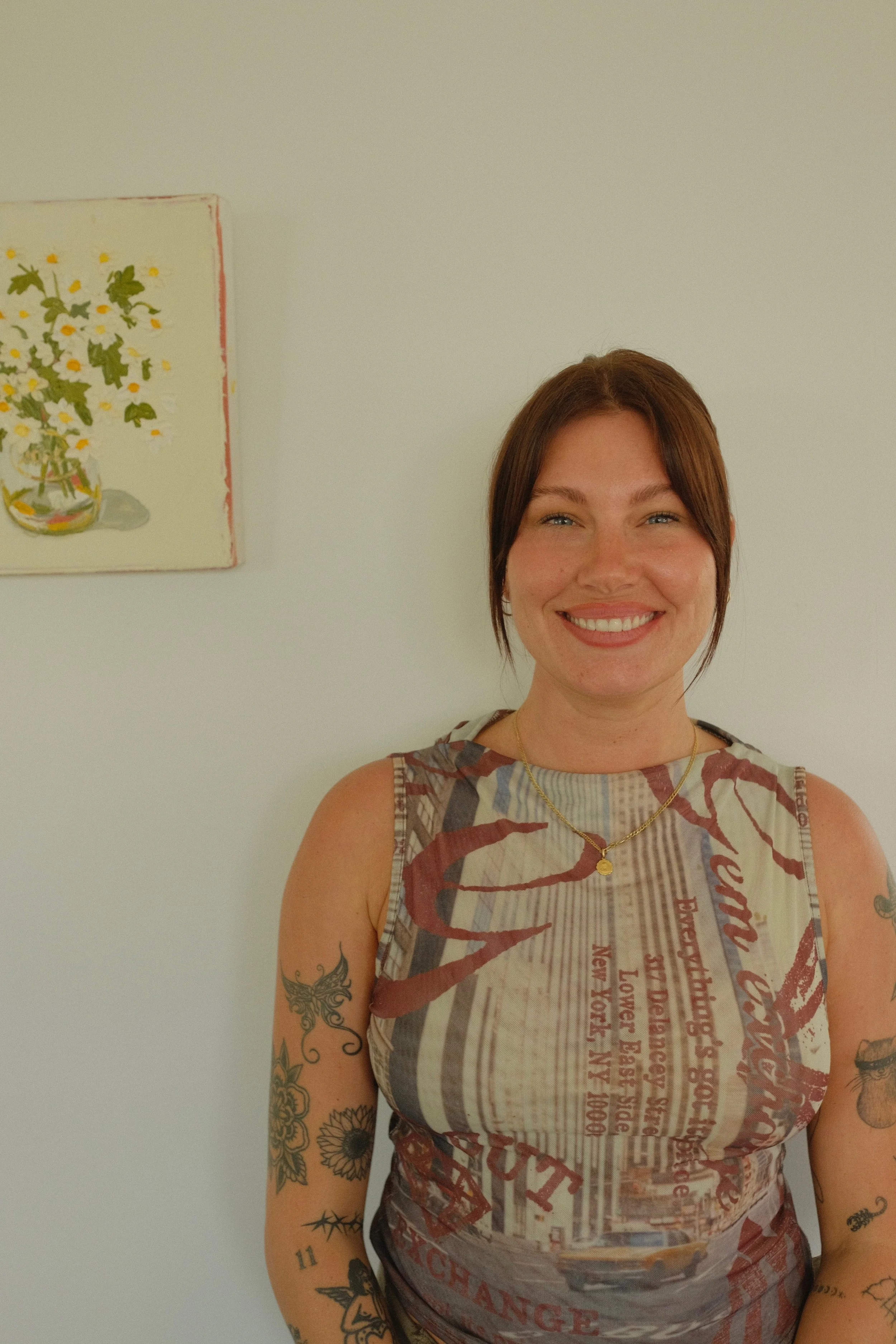 A smiling woman with brown hair, wearing a sleeveless graphic top and gold necklace, standing in front of a white wall with a floral painting hanging on it.