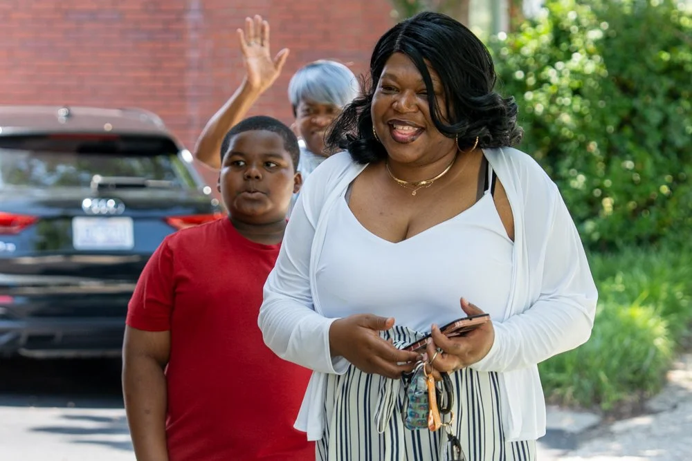 Three people outdoors, two women and a young boy, smiling and waving. The woman in the foreground has black hair and wears a white top and striped pants. The woman behind has short gray hair. The boy wears a red shirt. There is a black car and green foliage in the background.