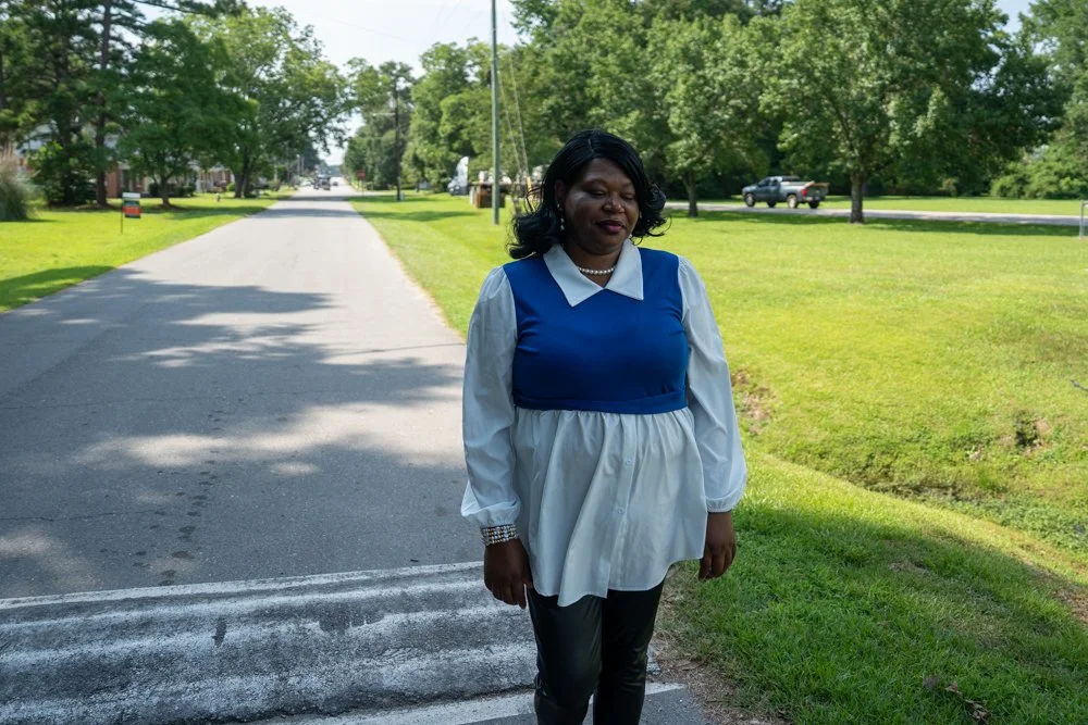 A woman standing on the side of a suburban street with green grass and trees, wearing a blue and white blouse with black pants.