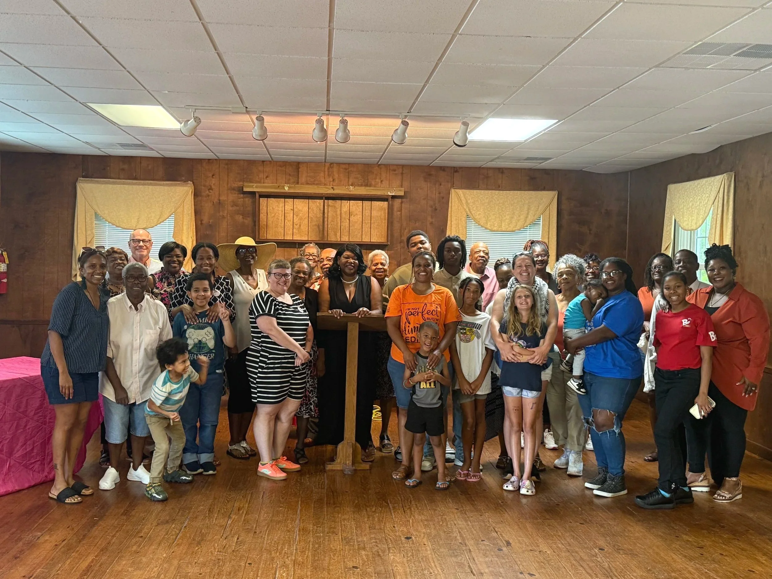 A large group of people, including adults and children, gathered in a room with wood paneling, yellow curtains, and a podium, posing for a group photo at an indoor event.
