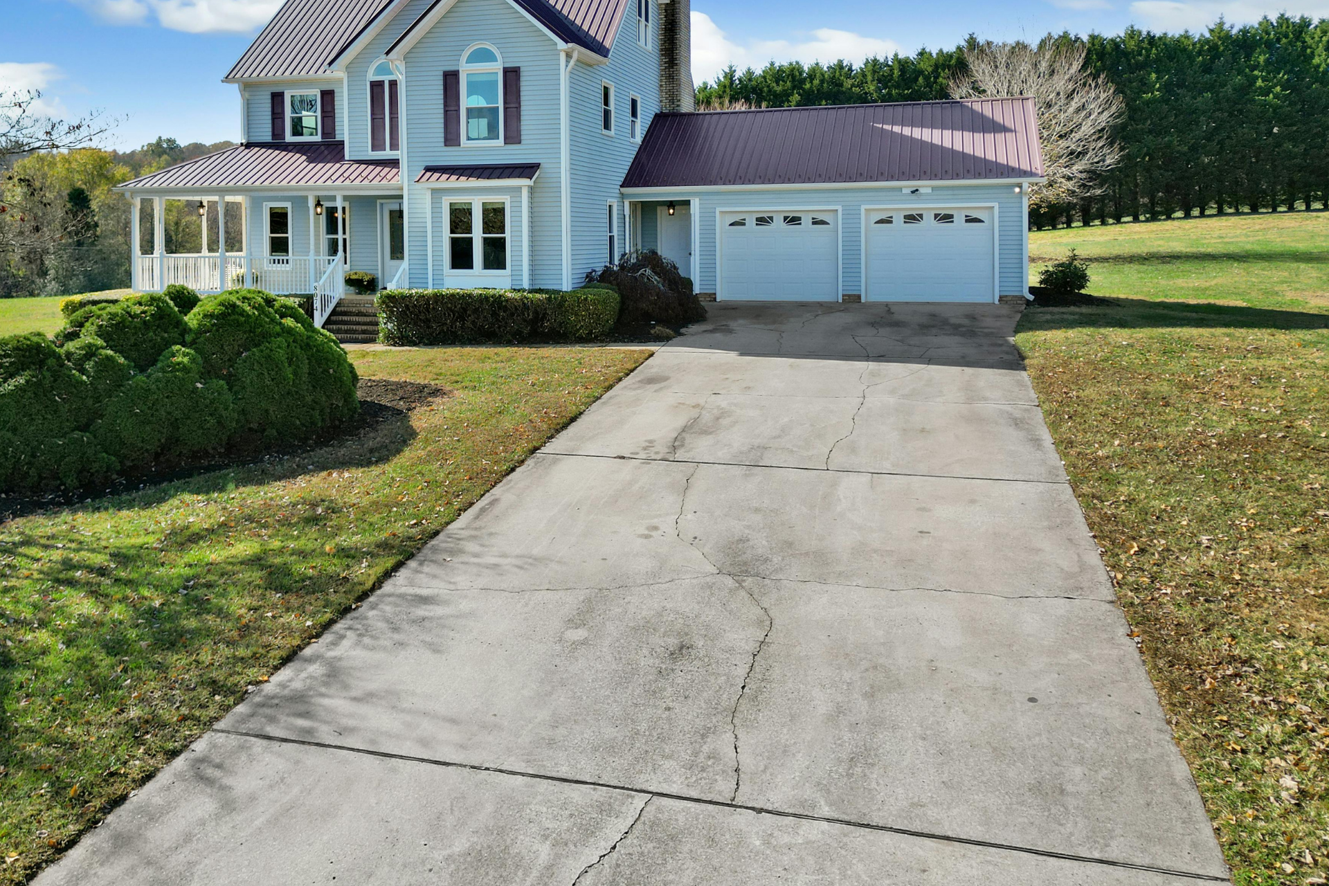 A large blue house with a metal roof, white trim, and a front porch with steps, surrounded by well-kept lawn and bushes, with a dirty concrete driveway leading to an attached garage.