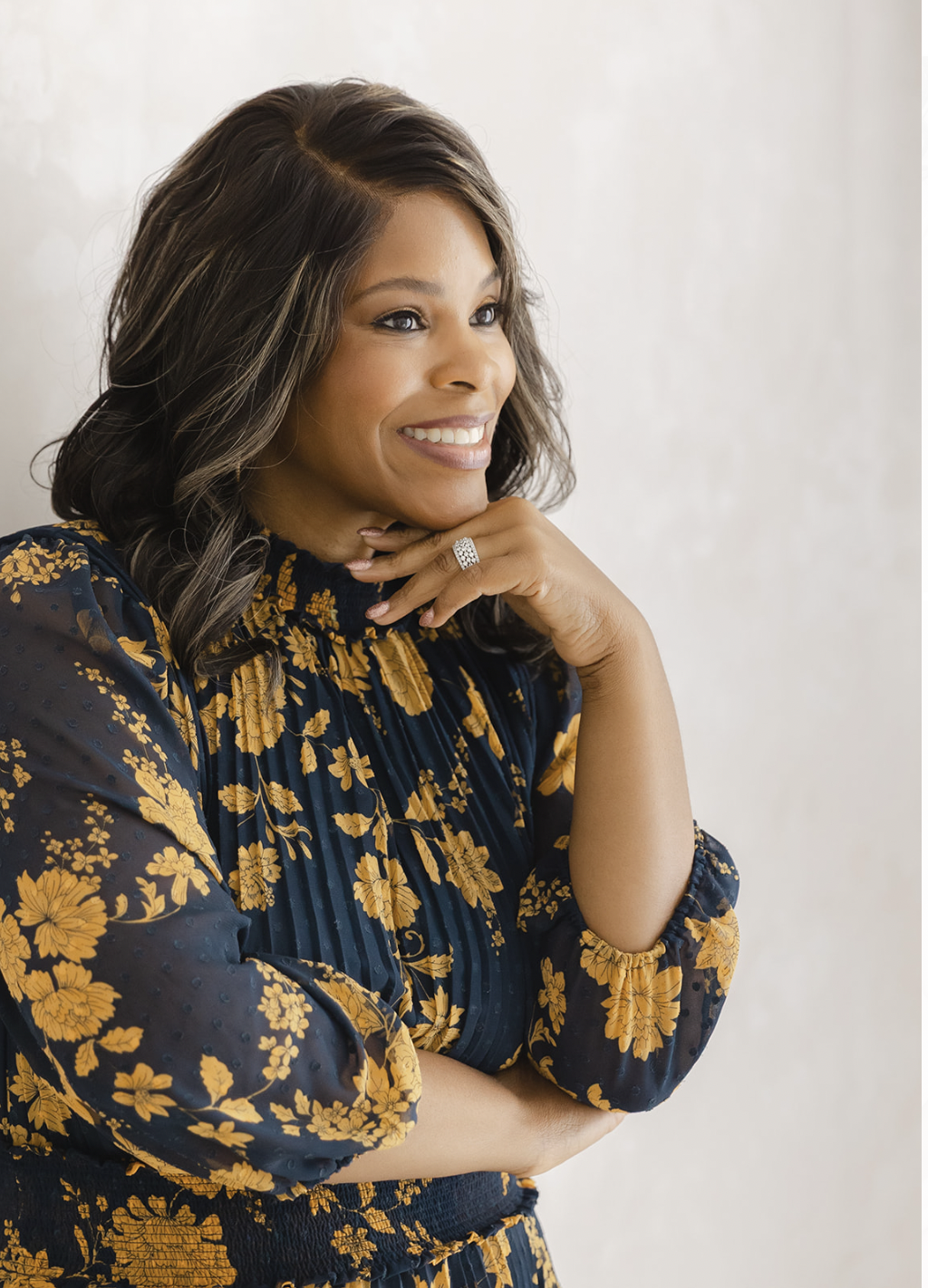 A woman with shoulder-length wavy hair, wearing a navy blue floral dress with yellow flowers, smiling and resting her chin on her hand, with a plain light-colored background.