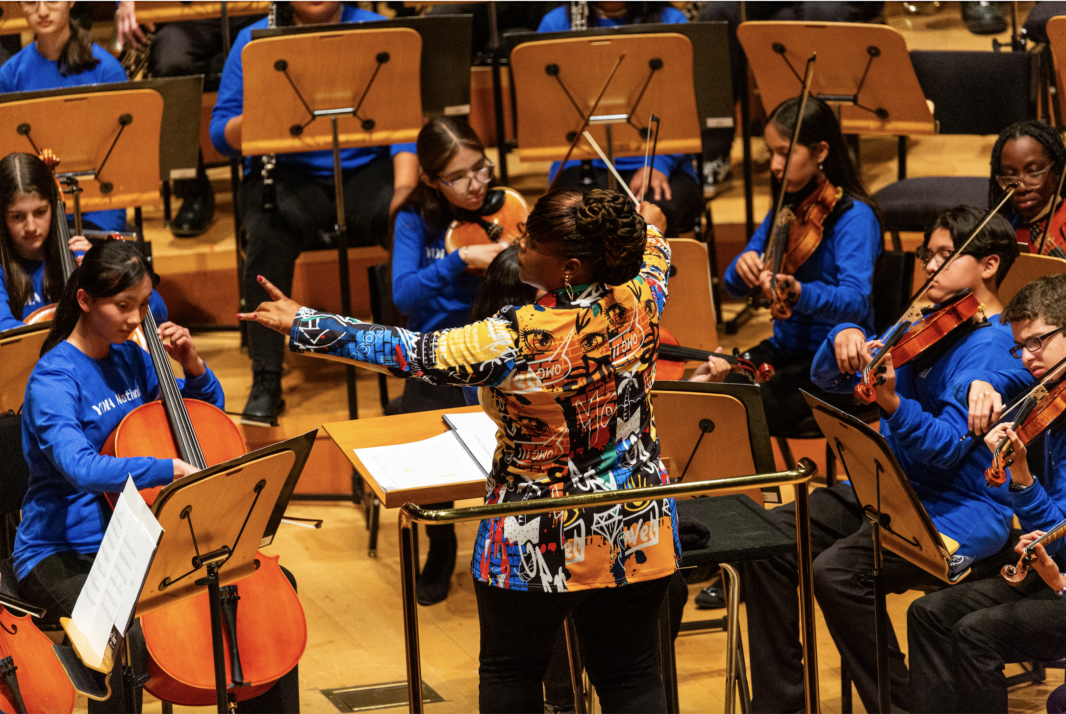 A symphony conductor with a colorful jacket leads a youth orchestra to young musicians playing violins and cellos, all wearing blue shirts in a concert hall.