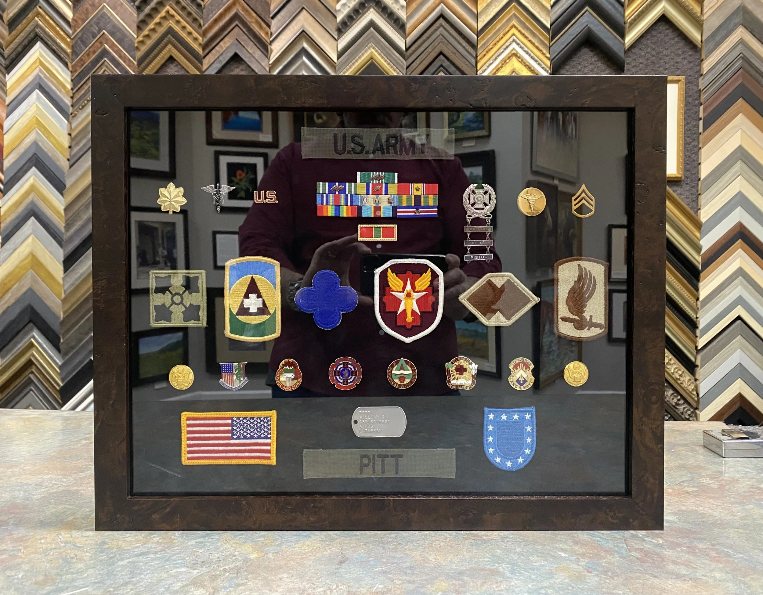 A mirror displaying various military patches, ribbons, and badges inside a frame labeled "U.S. ARMY" and "PITT" with a reflection of a person taking the photo.