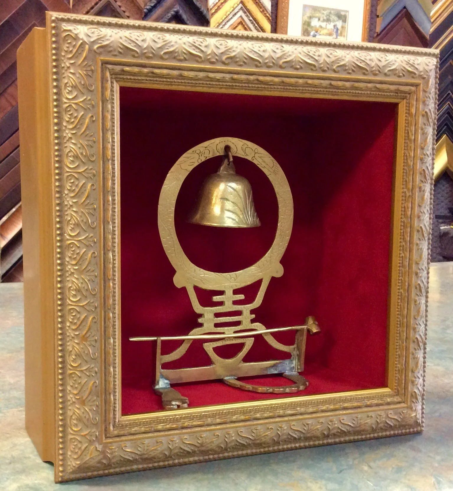 A gold bell inside a decorated wooden display case with a red velvet background, featuring intricate gold and silver detailing and a metallic stand at the base.