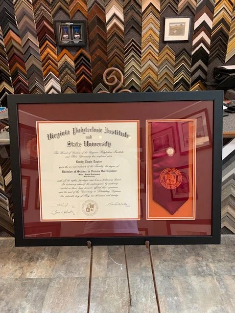 A framed diploma from Virginia Polytechnic Institute and State University, along with a medal or medallion in a protective case, displayed on a stand in a framing shop with various frame samples on the wall.