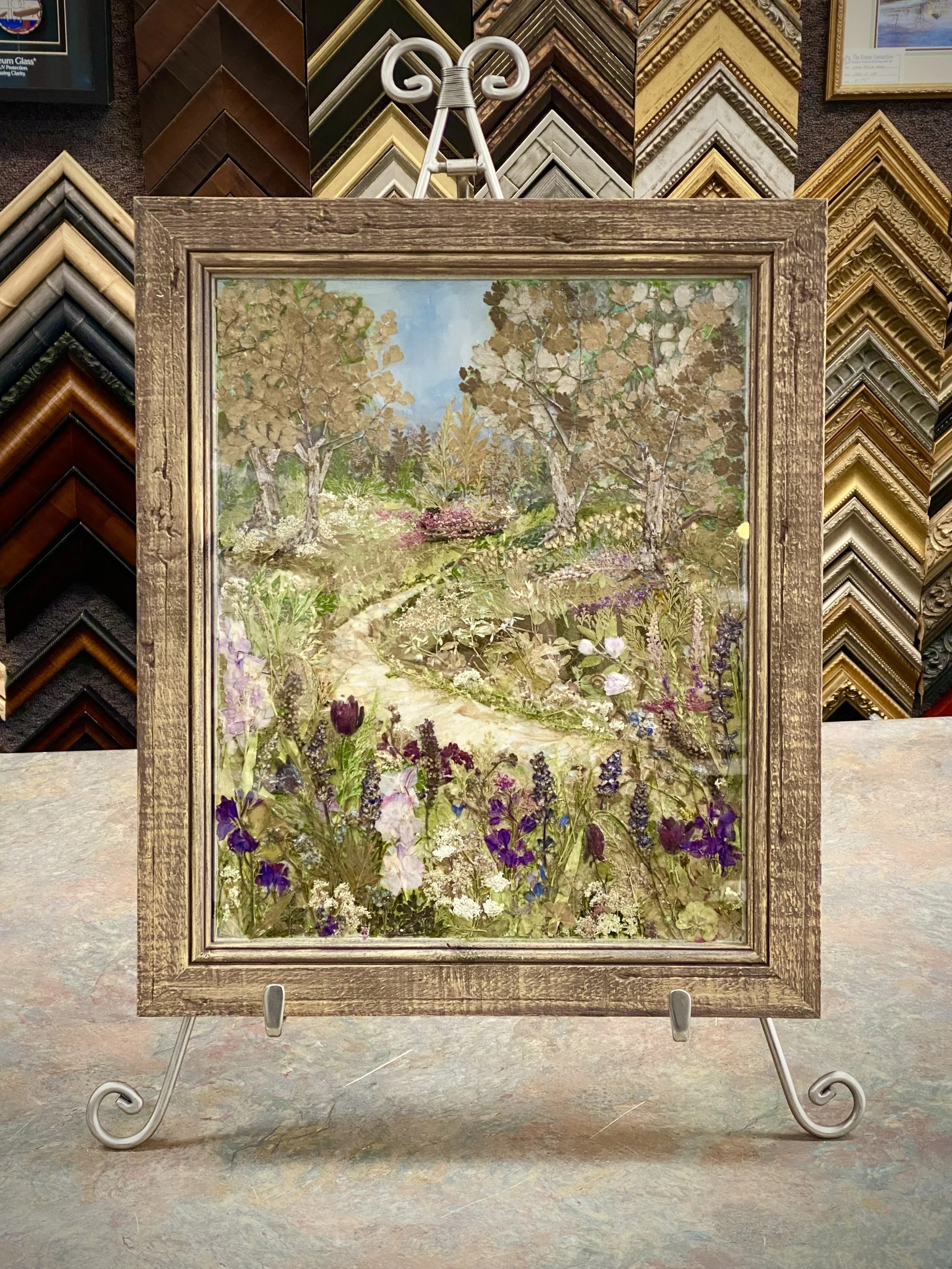 Framed landscape painting of a garden with trees and colorful flowers, displayed on a stand in an art store surrounded by multiple picture frame samples.