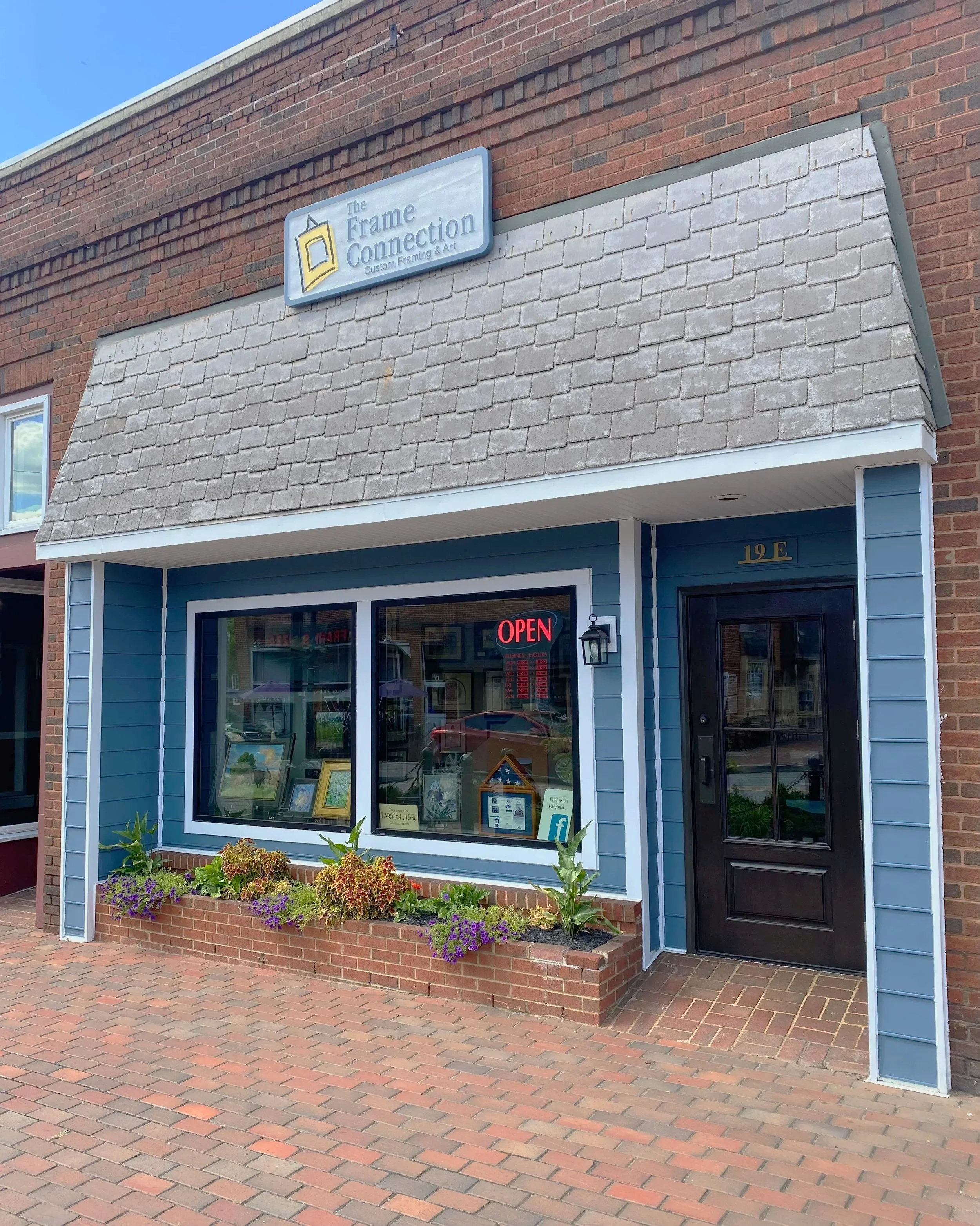 Exterior view of a storefront called "The Frame Connection" with a blue facade, black door, and large window displaying artwork. An illuminated red "OPEN" sign is in the window. The building has a brick sidewalk in front and a flower bed with green p