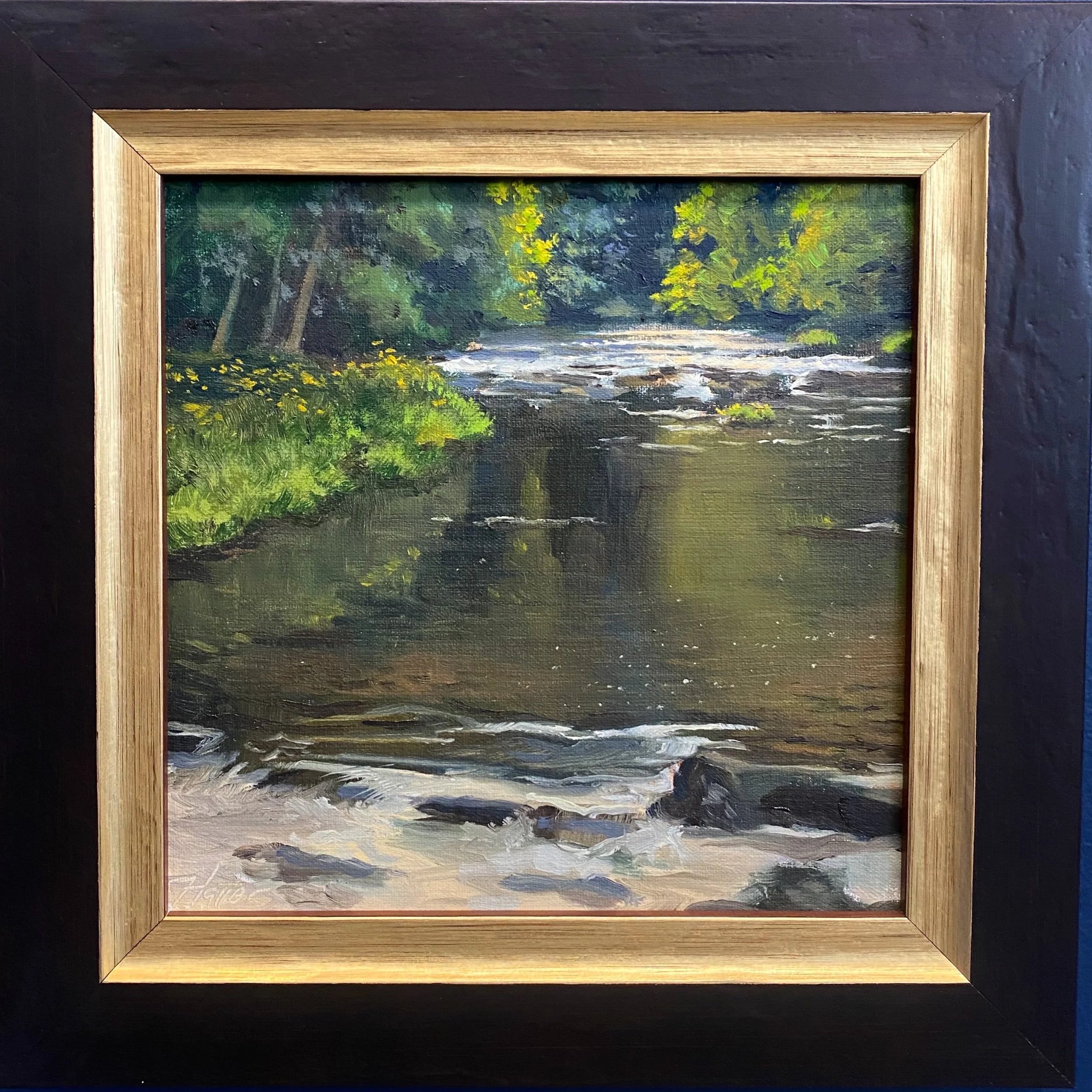 A framed landscape oil painting of a river surrounded by green trees and yellow flowers, with a dark wooden frame, set against a black background.