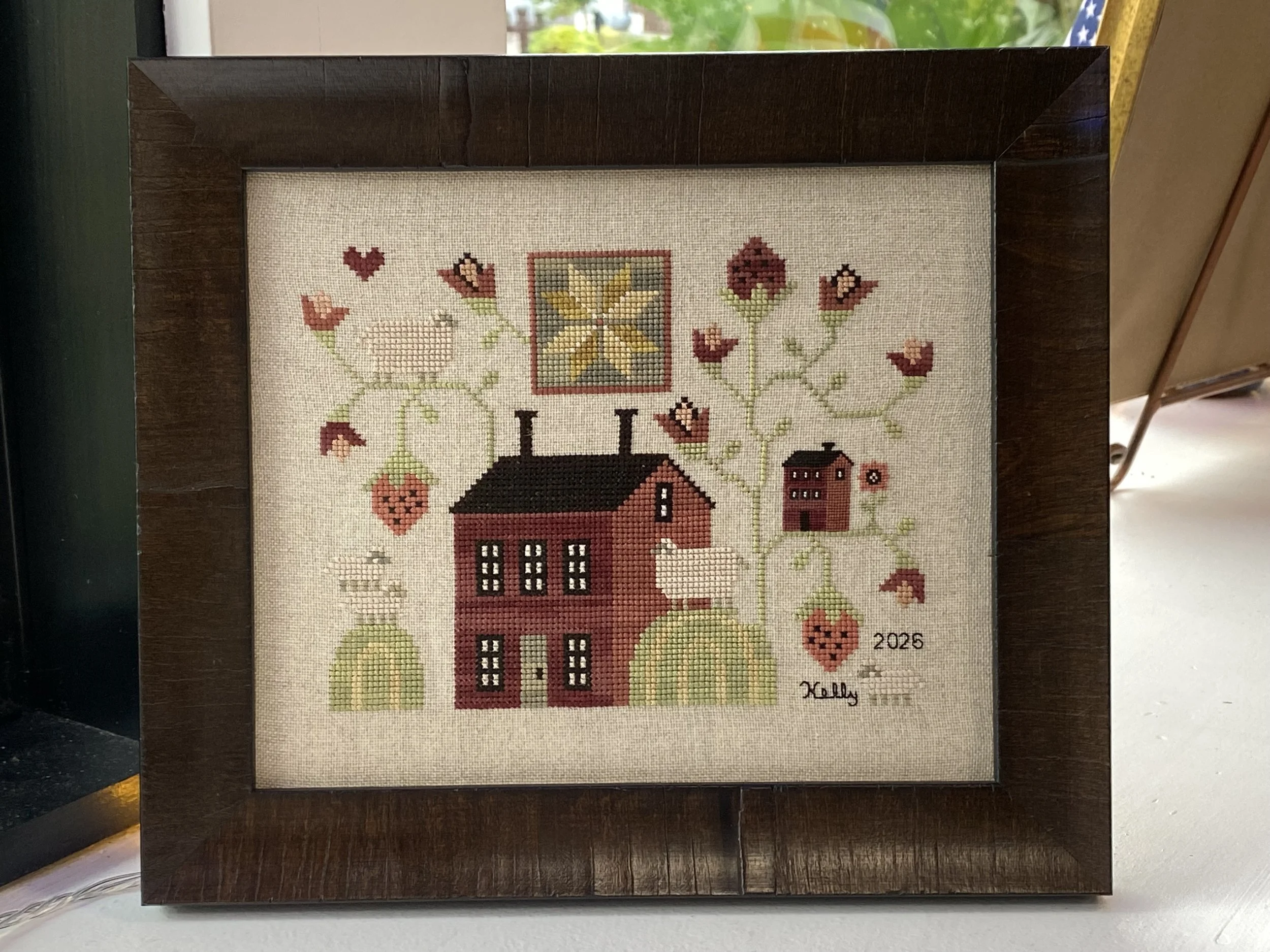 Cross-stitch artwork framed in a dark wood frame, featuring a red barn, trees, and floral patterns, with the year 2025 and the signature 'Kell'.