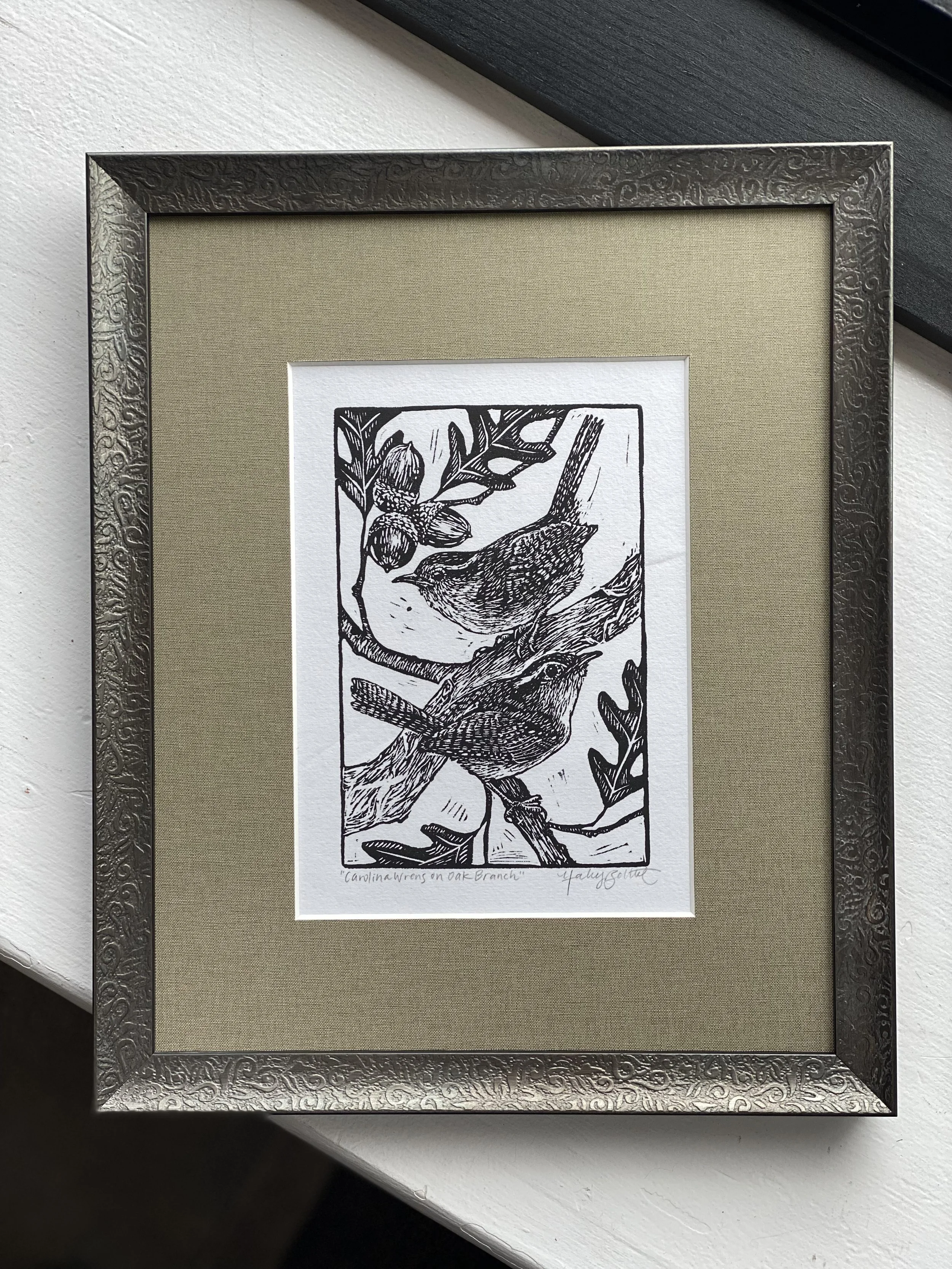 A black and white drawing of a bird perched on an oak branch with acorns, framed with a decorative metal frame and beige mat, titled 'Canolina Wrens on Oak Branch'.