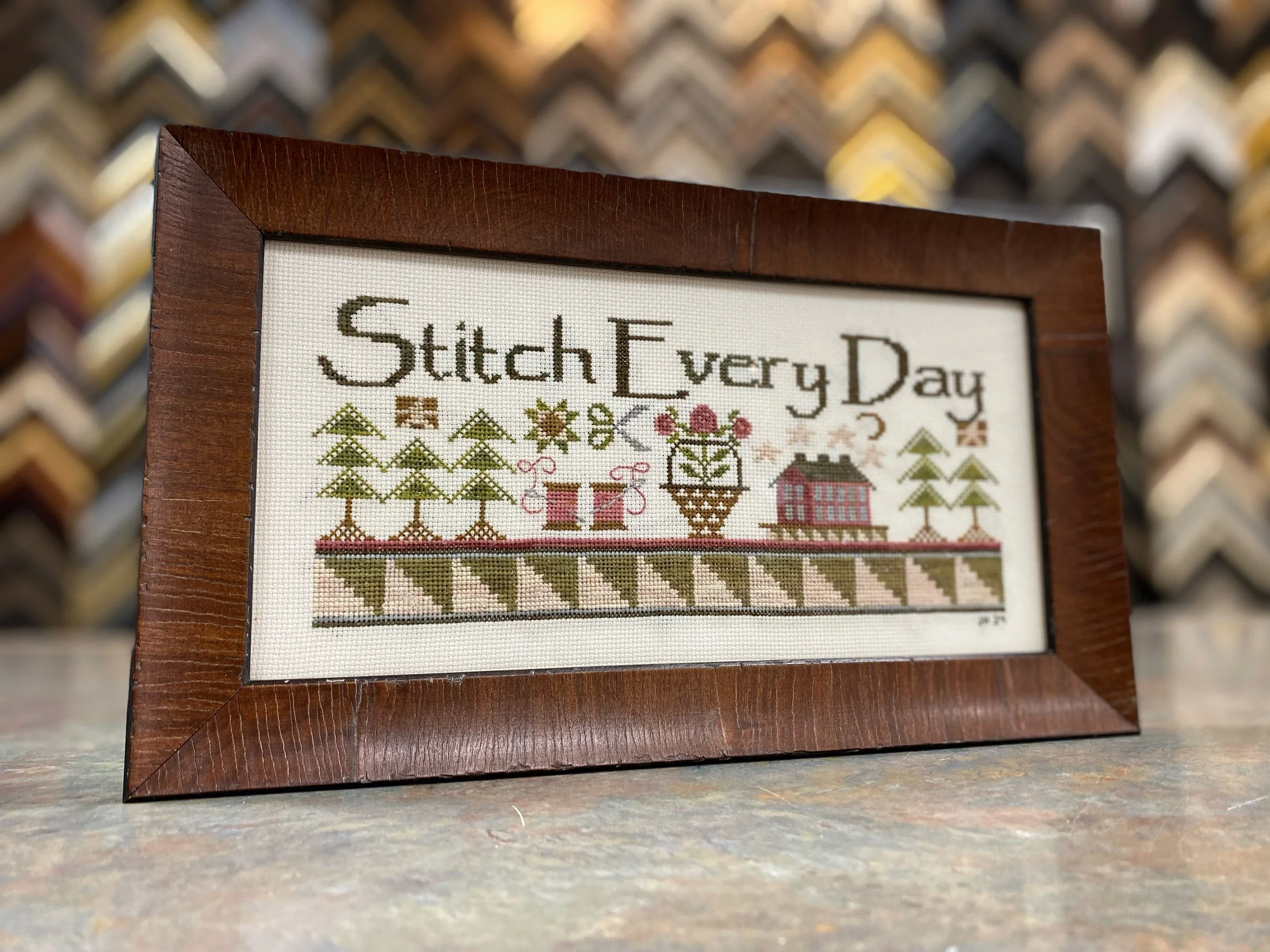 Cross-stitch embroidery with the phrase "Stitch Every Day" and decorative elements including trees, stars, and a house, framed in dark brown wood.