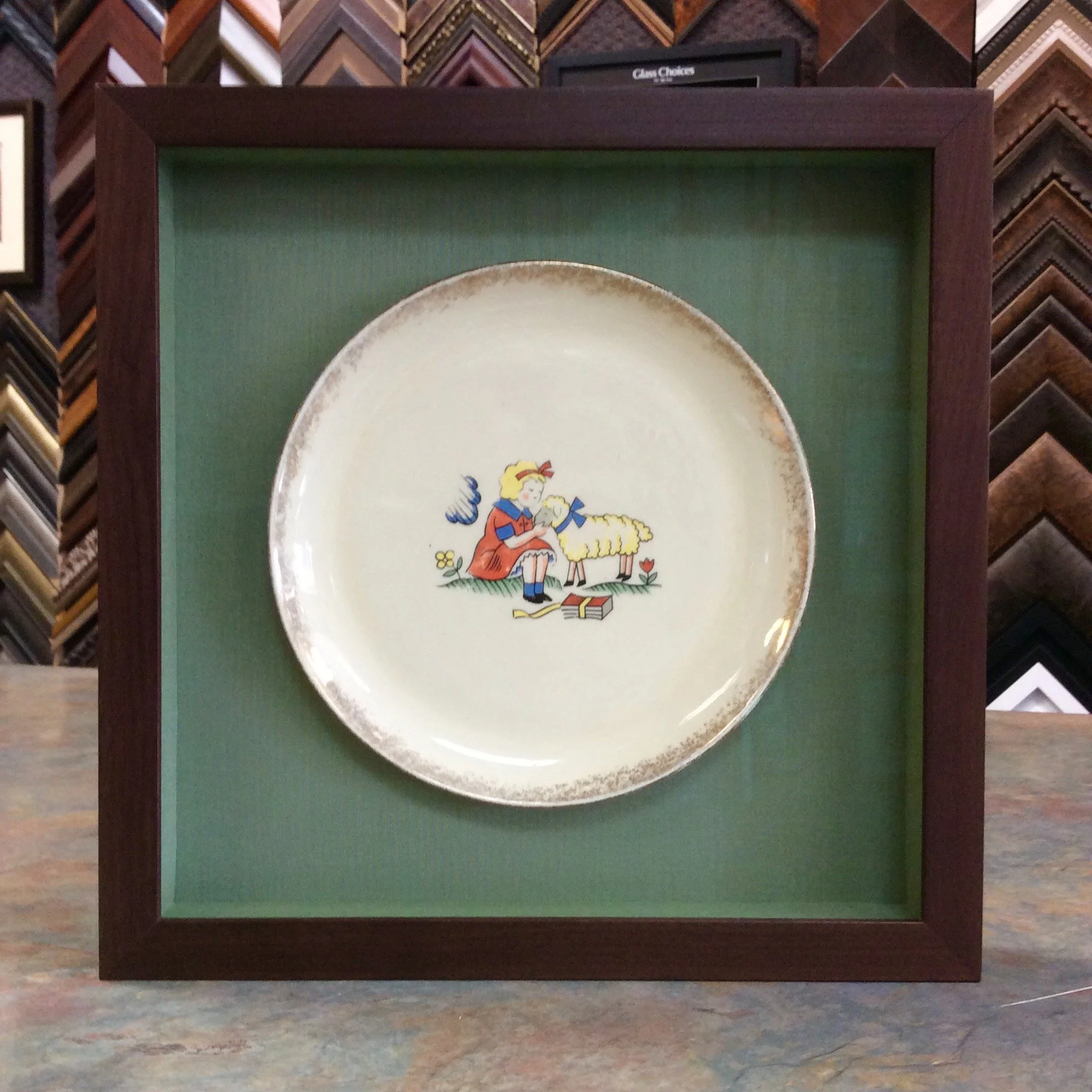 Framed ceramic plate with a colorful illustration of a girl feeding a sheep, placed on a green background inside a wooden frame.