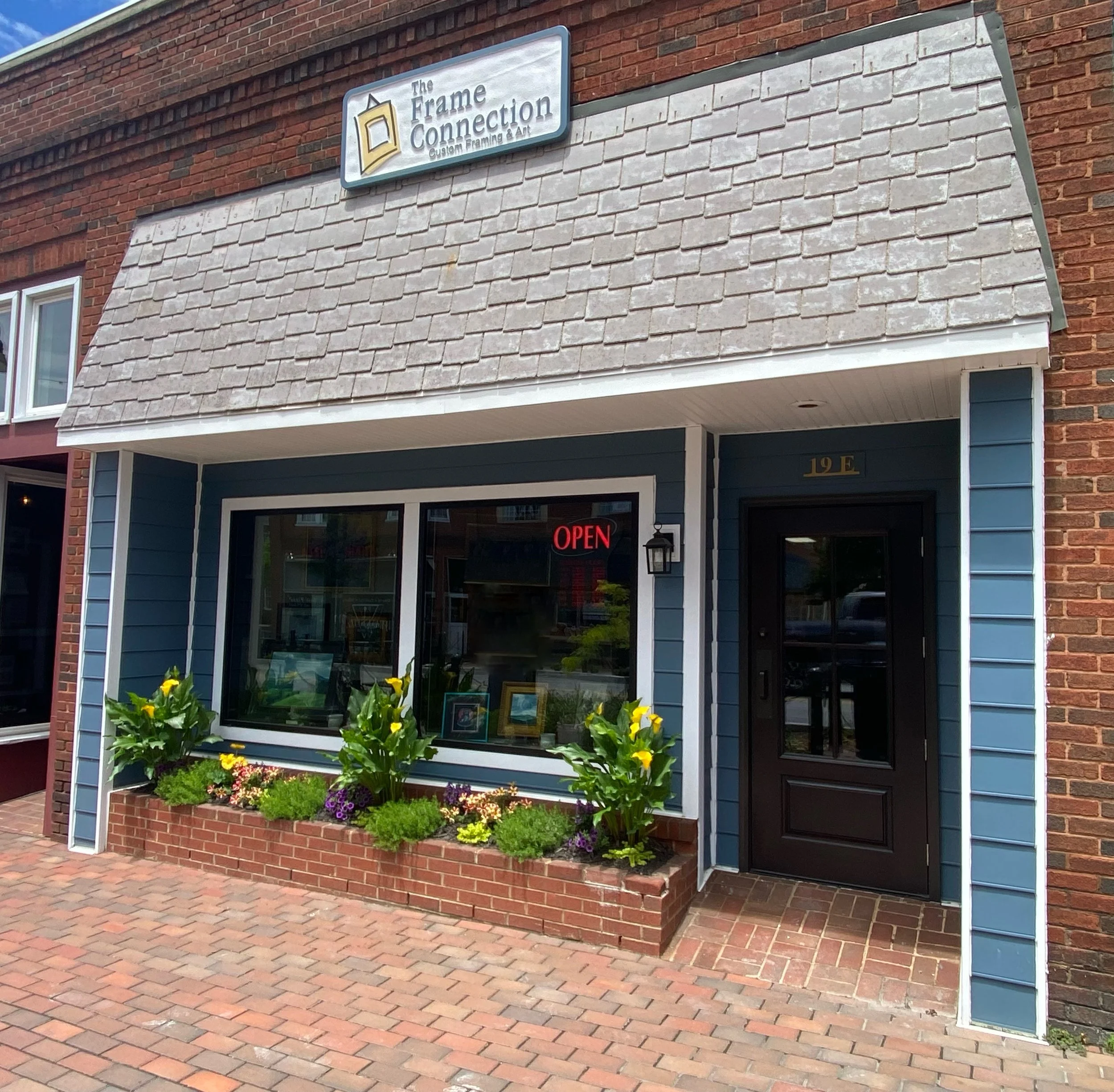 Storefront with a sign reading 'The Frame Connection, Custom Framing & Art'. The building has red brick and blue siding, with a window display of artwork and a flower bed in front.
