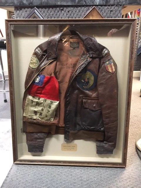 A framed display case with a vintage brown leather military jacket, decorated with patches and insignia, including a blue and red flag patch on the sleeve, and a cloth tag or patch inside the jacket.