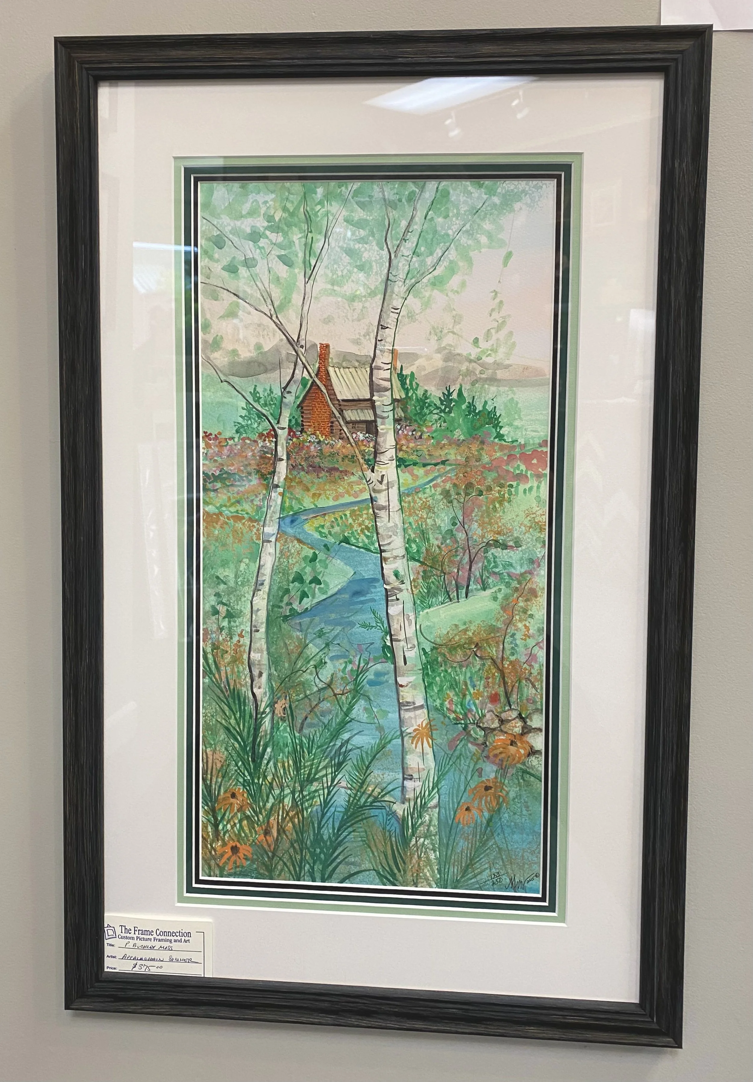 Framed watercolor painting of a landscape with tall trees, a winding creek, colorful bushes, and a small house in the distance.