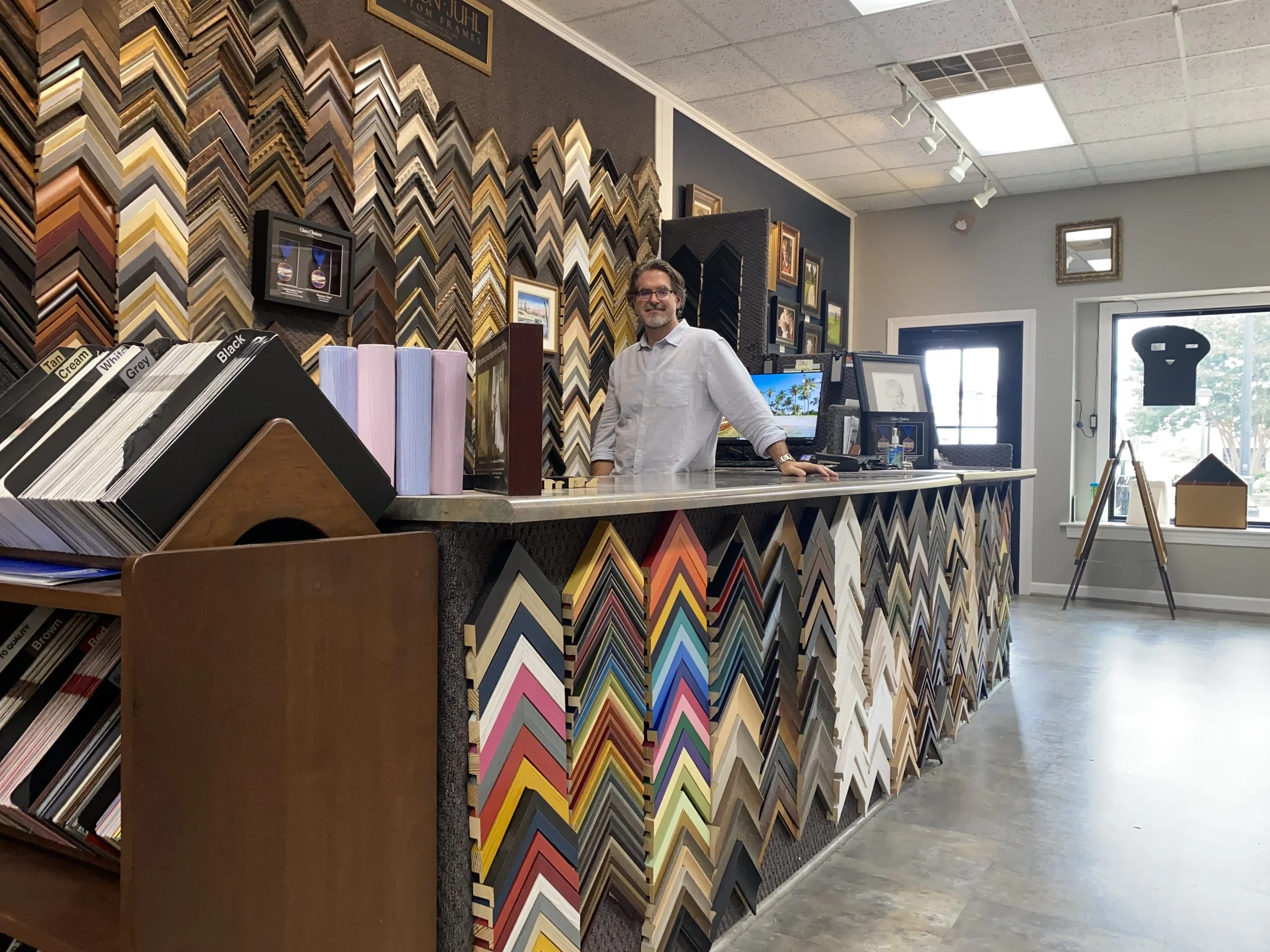 A man behind a counter in a store that sells picture frame moldings, with shelves full of various frame samples on the wall and a counter with color samples and framed artwork.