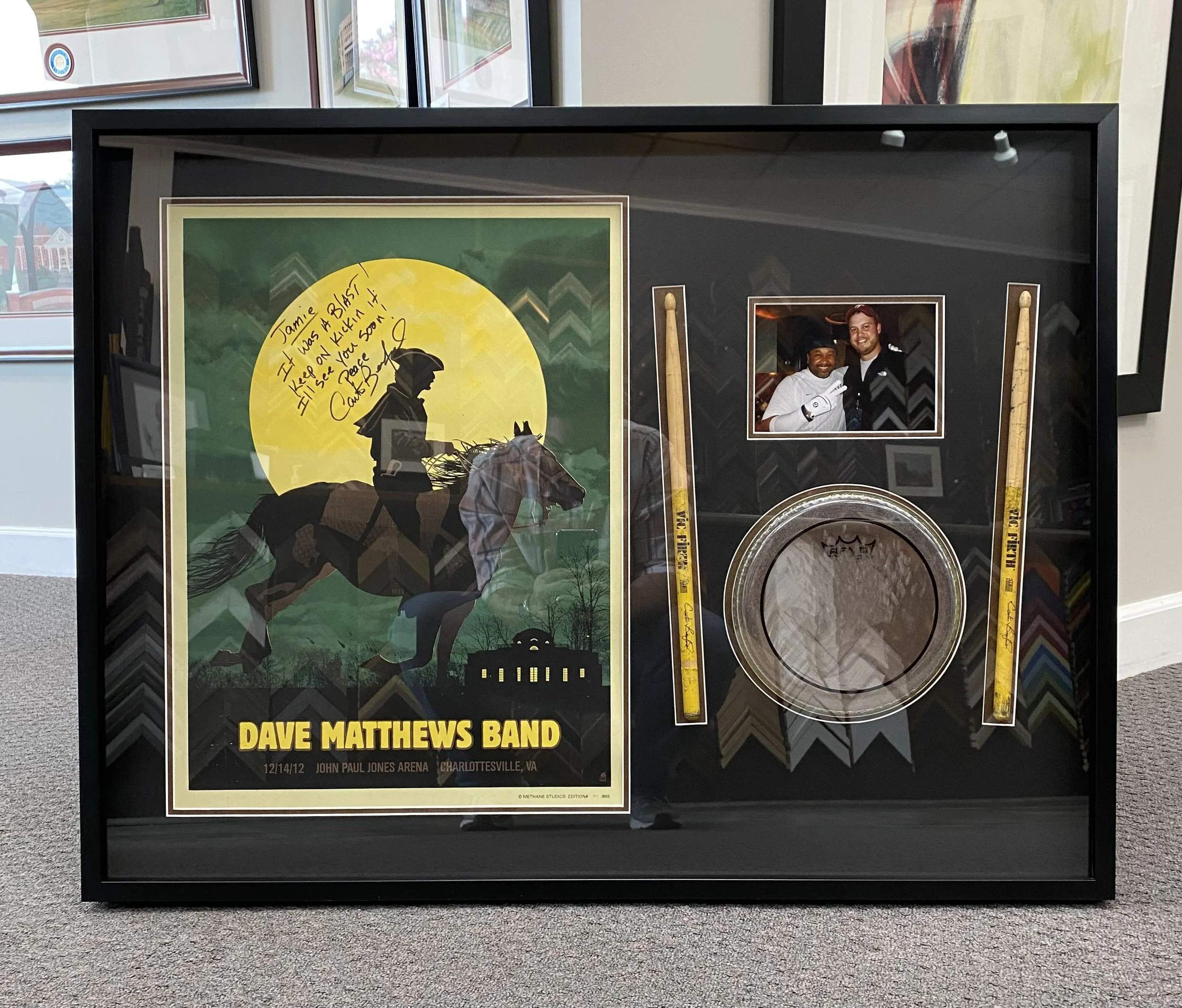 A framed concert poster for Dave Matthews Band at John Paul Jones Arena, Charlottesville, VA, on December 14, 2012, with a window reflection and two yellow caution tape strips.
