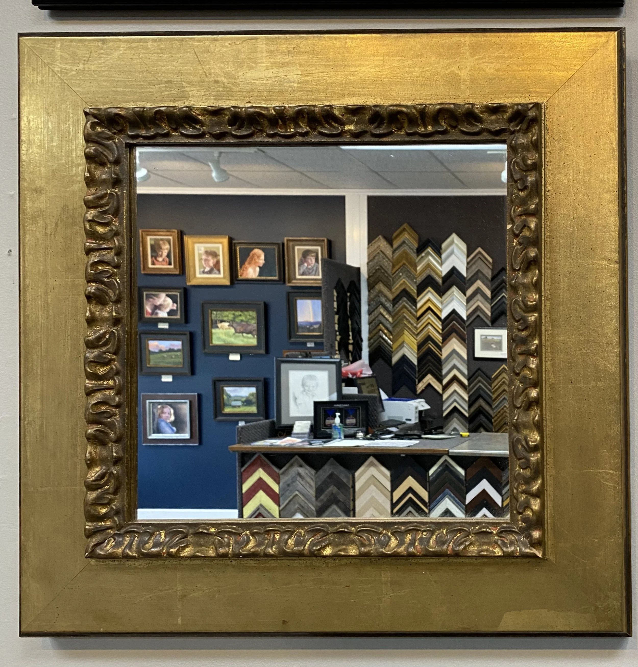 A decorated mirror with a gold frame, hanging on a wall. Reflected in the mirror is a dark blue wall adorned with various framed pictures and artwork, some of which depict nature and people. Expert custom mirror framing in Salem, VA, and near Roanoke