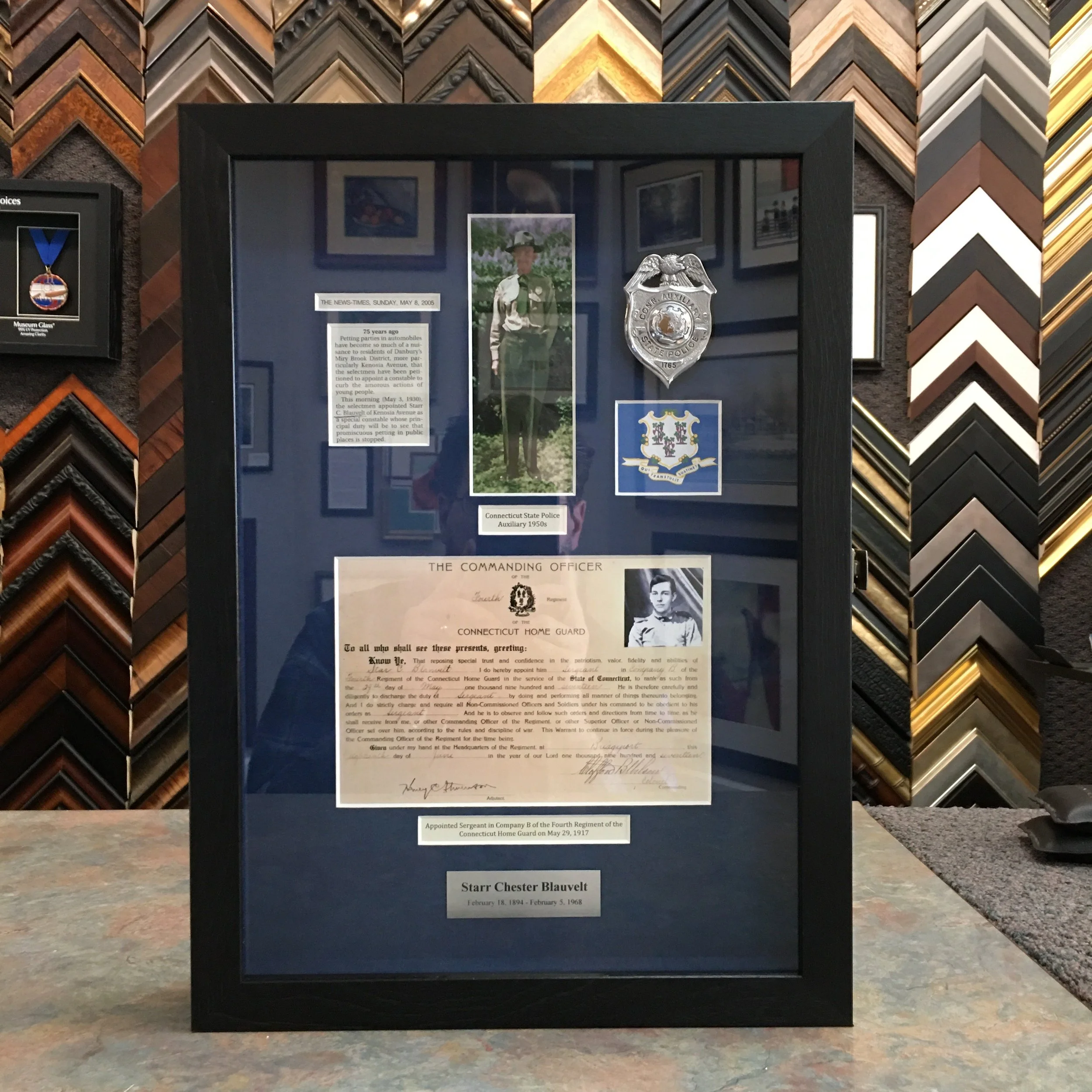 Display case containing historical documents and photographs related to the Connecticut State Police Auxiliary from the 1950s, with background samples of picture frame moulding.
