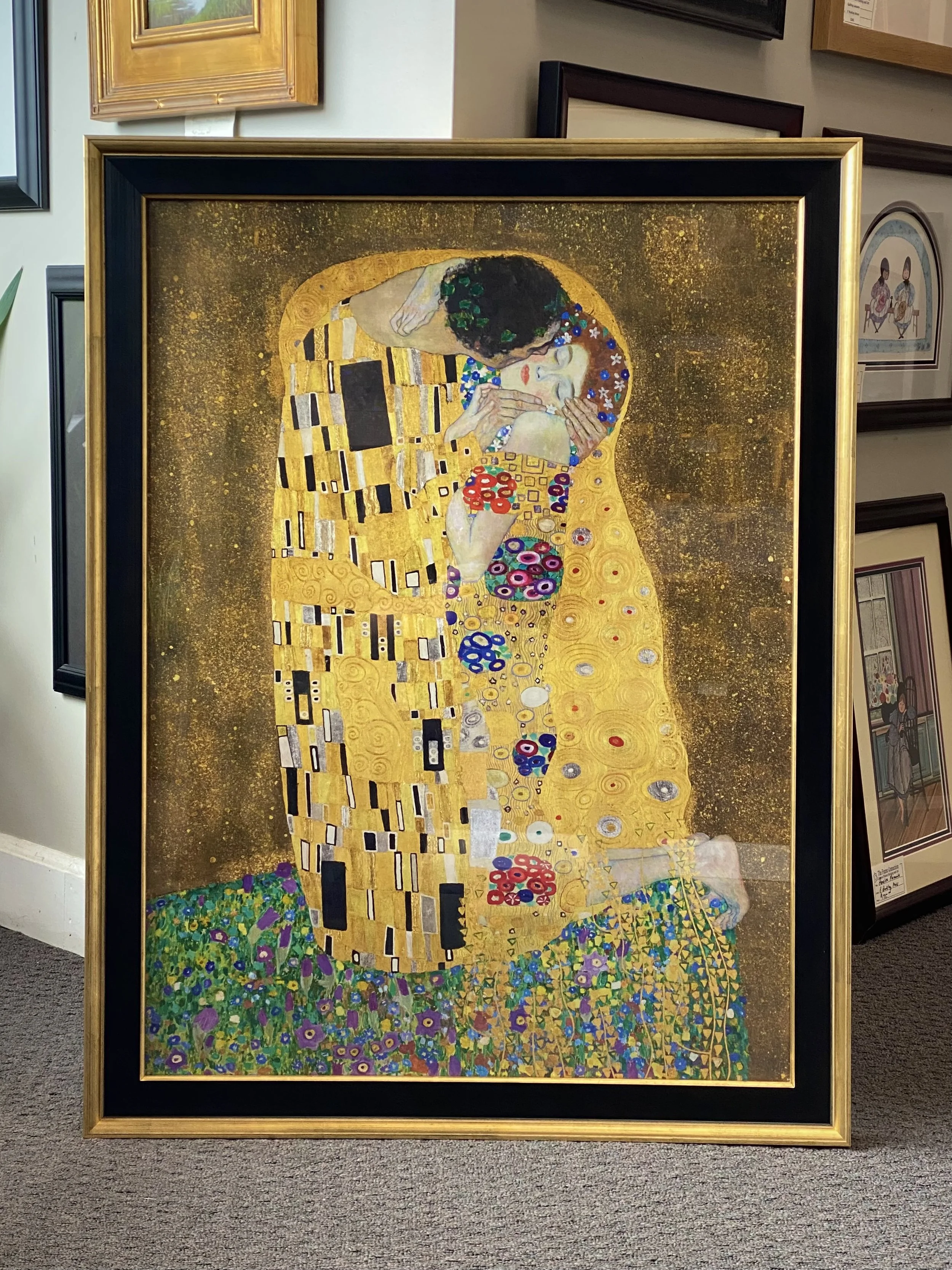 A framed reproduction of Gustav Klimt's painting 'The Kiss' displayed on a wall with other framed artworks around it. The painting depicts a couple embracing, with intricate gold patterns and floral motifs.