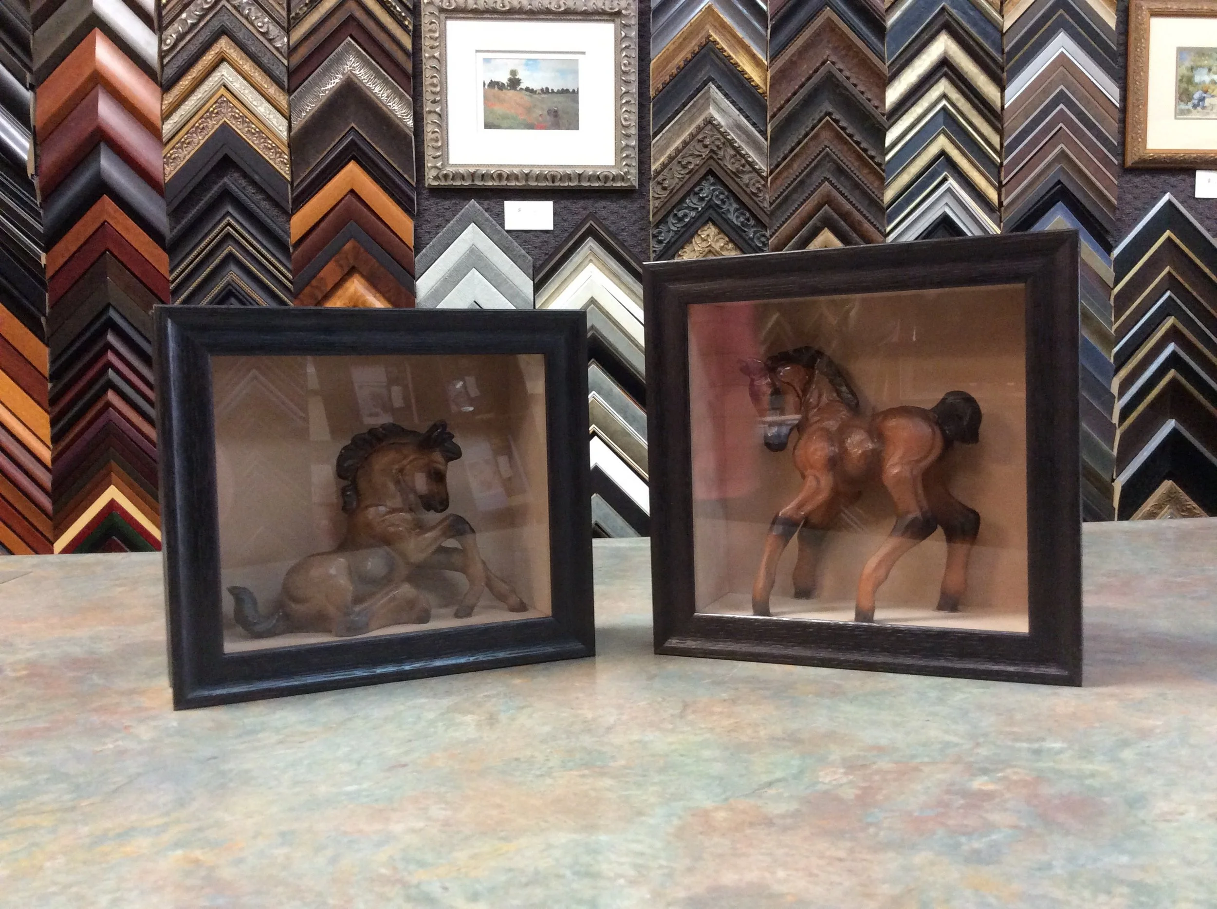 Two glass display cases containing horse sculptures, with a background of a wall decorated with various colorful picture frame moldings and framed artwork.