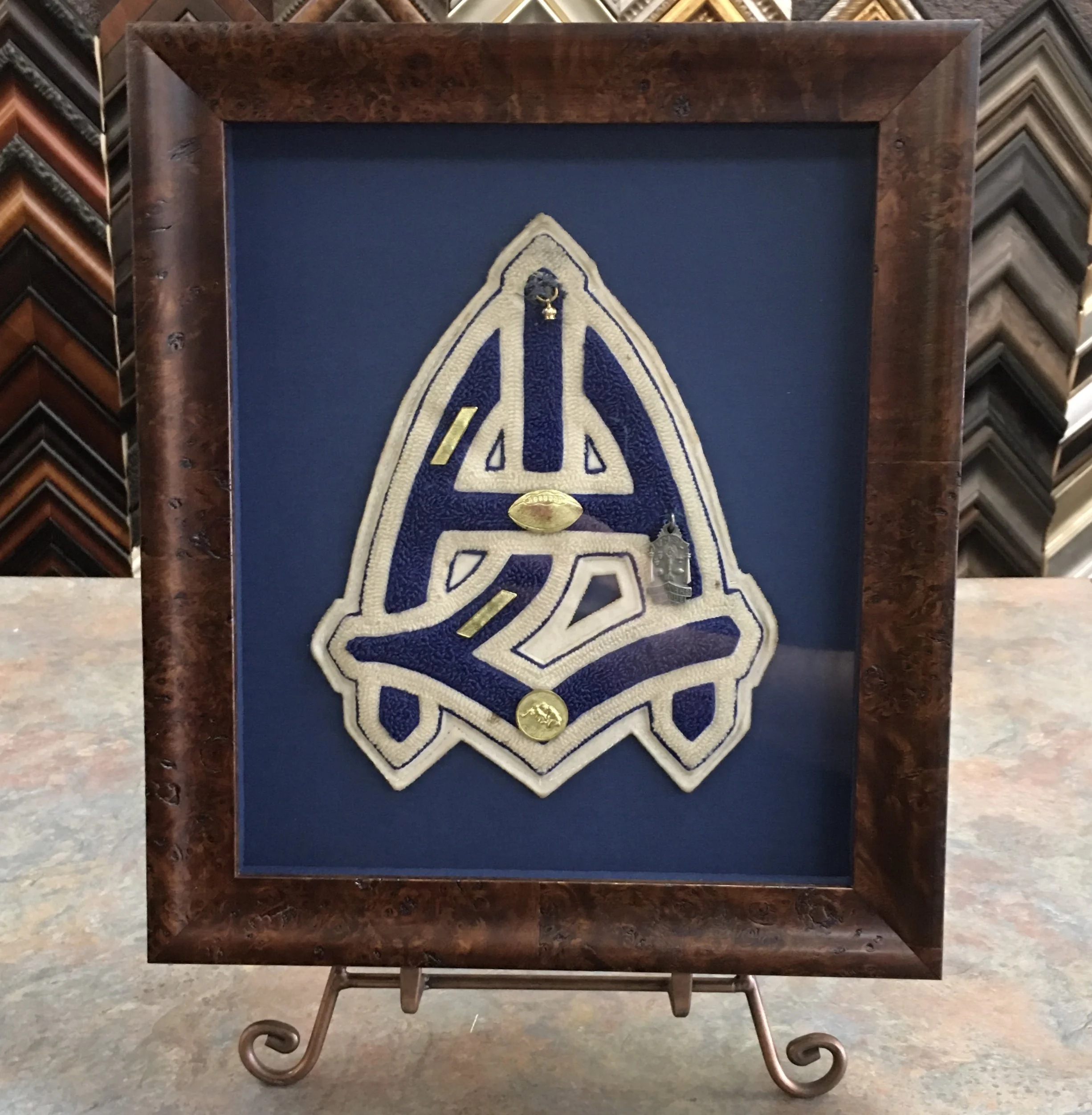 A blue framed embroidered patch with a gold ring and a small metallic badge inside, set on a textured wooden table with a variety of picture frame samples in the background.