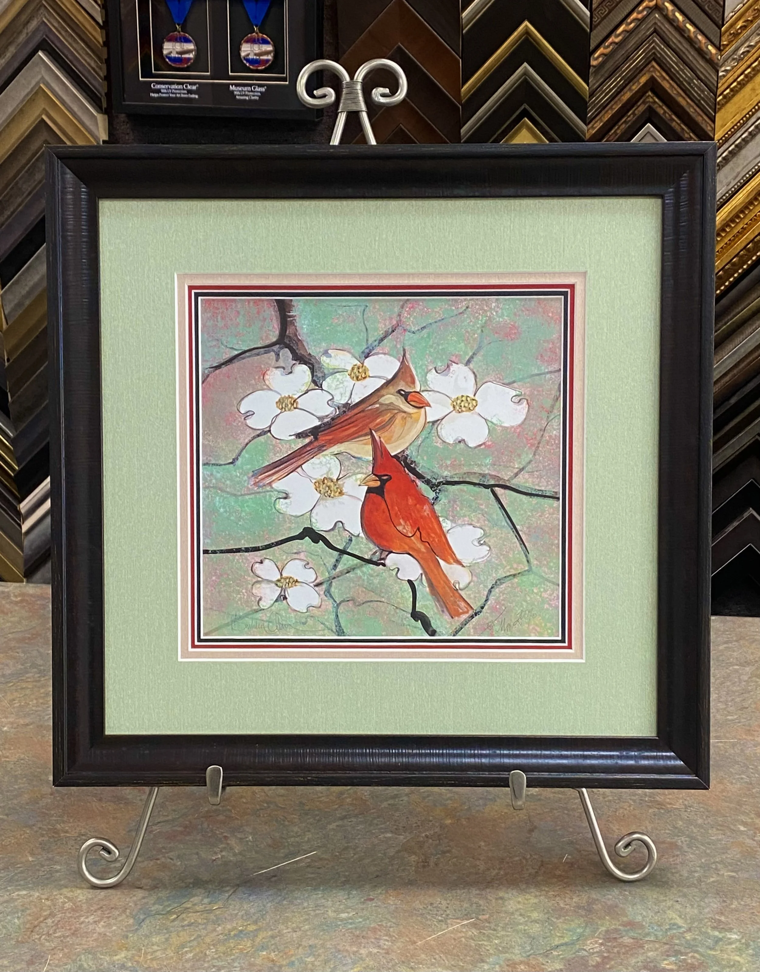 Framed artwork of two cardinals sitting on a branch with white blossoms, with a light green mat and gallery style frame, displayed on a stand in an art store.