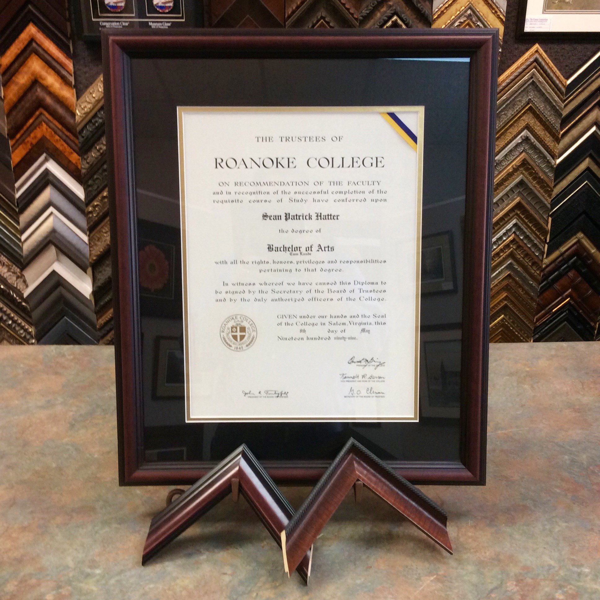 Certificate framed with a dark wooden border, mounted on a display wall showcasing various picture frame samples in different styles and finishes.