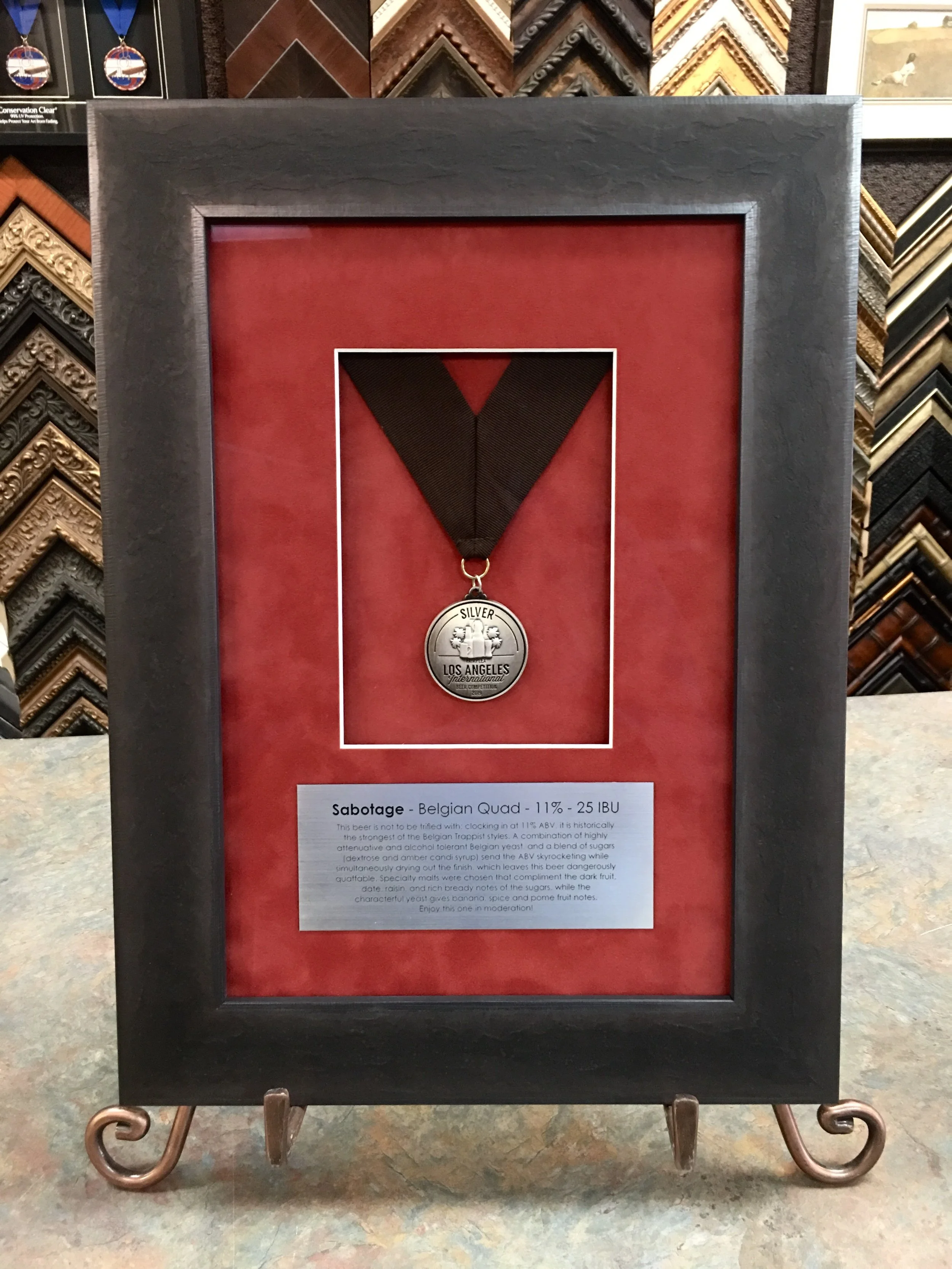 A framed beer medal with a black ribbon and a silver medallion labeled 'SILVER LOS ANGELES International'. The background is red, and there is a description and information about the beer 'Sabotage - Belgian Quad' below the medal, including its alcoh