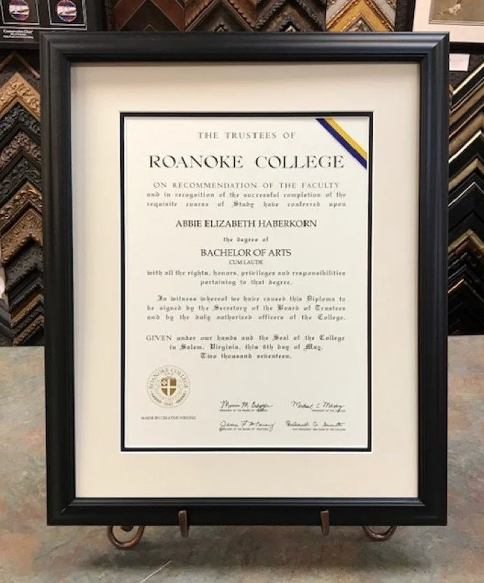 A framed diploma from Roanoke College awarded to Abbie Elizabeth HaberKorn for earning a Bachelor of Arts degree, signed by college officials, displayed on a stand in a store with various picture framings in the background.