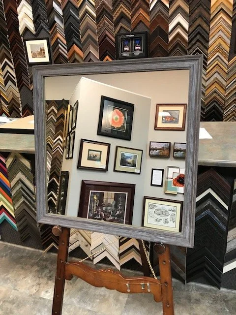 A large rectangular mirror on a wooden stand reflects a display of framed artwork and photographs on a white wall, with a wall of various picture frame samples in the background. Expert custom mirror framing in Salem, VA, and near Roanoke, VA.
