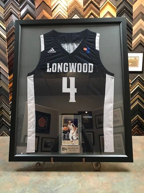 Framed basketball jersey with the word 'LONGWOOD' and the number 4, displayed behind glass. The jersey is black with white accents, and there is a small photograph or card below the jersey in the frame.