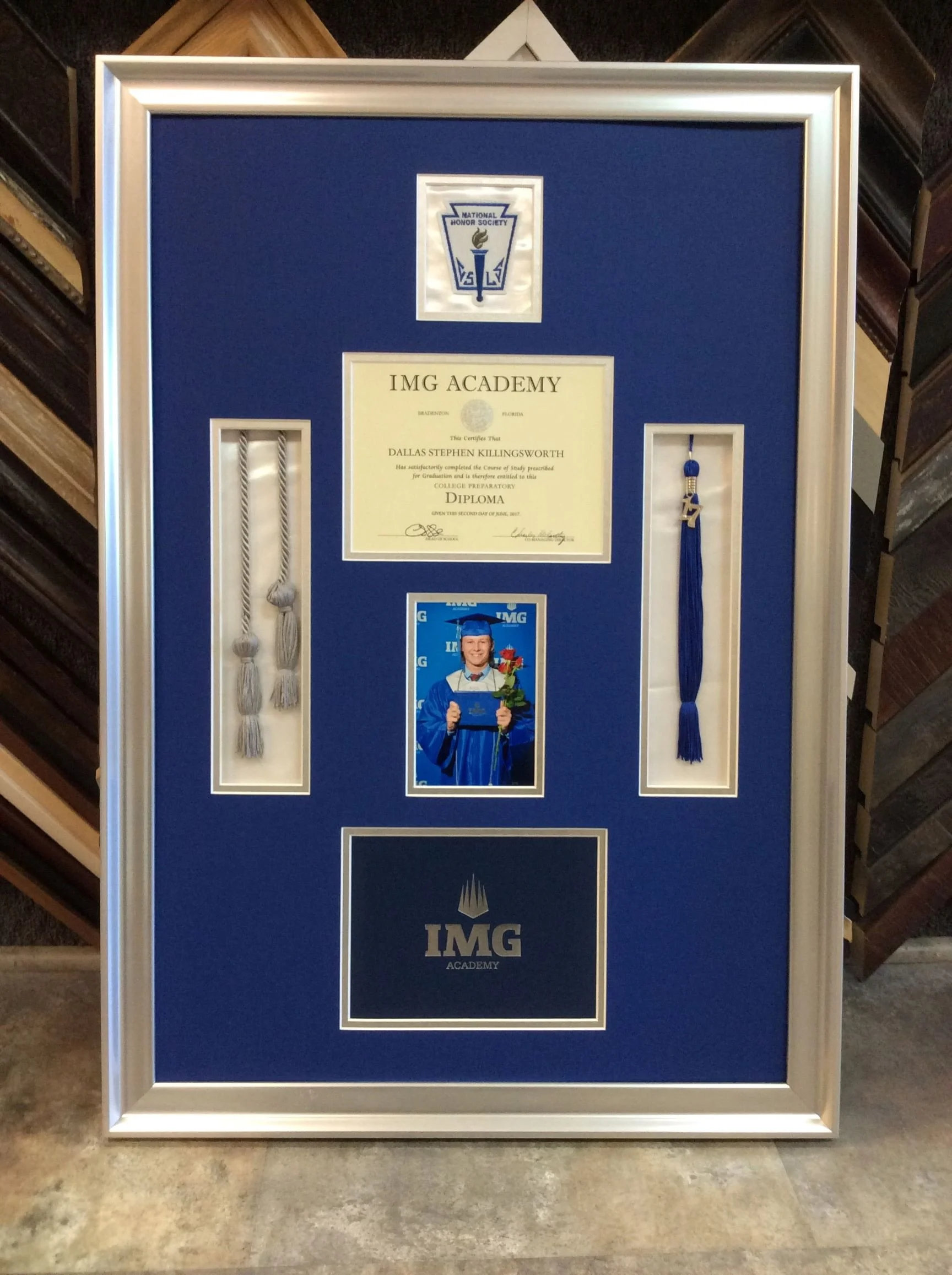 A framed display with a blue background containing various items associated with education and achievement. At the top, there is a logo of the National Honor Society. In the center, there is a diploma from IMG Academy awarded to Dallas Stephen Killin