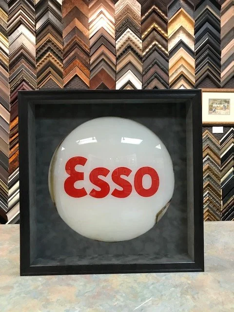 Framed Esso sign featuring a white globe with red Esso logo, placed on a table in front of a display of various picture frame samples.