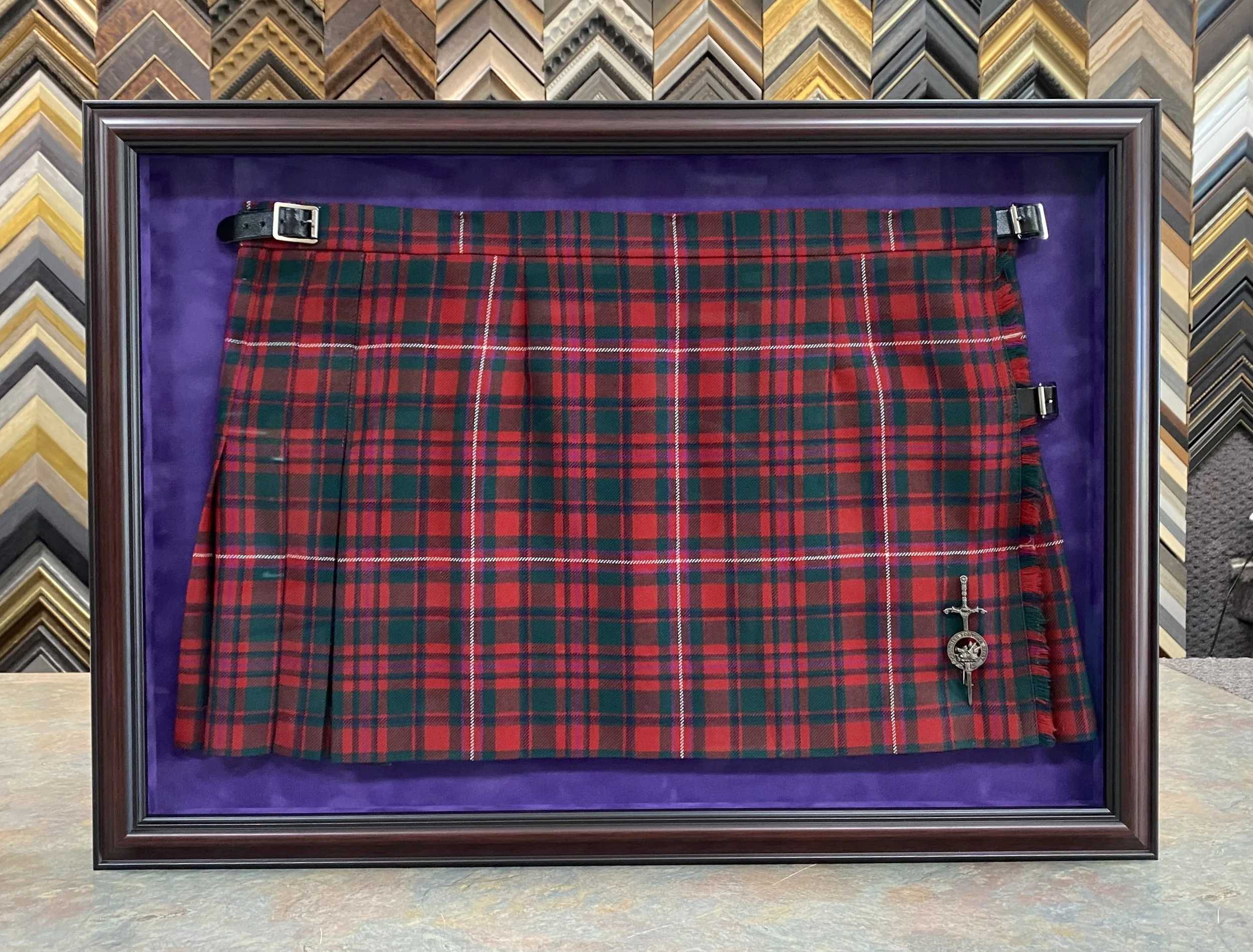 A framed tartan plaid fabric with red, green, and white pattern, displayed against a purple background in a store with a background of various picture frame moldings.