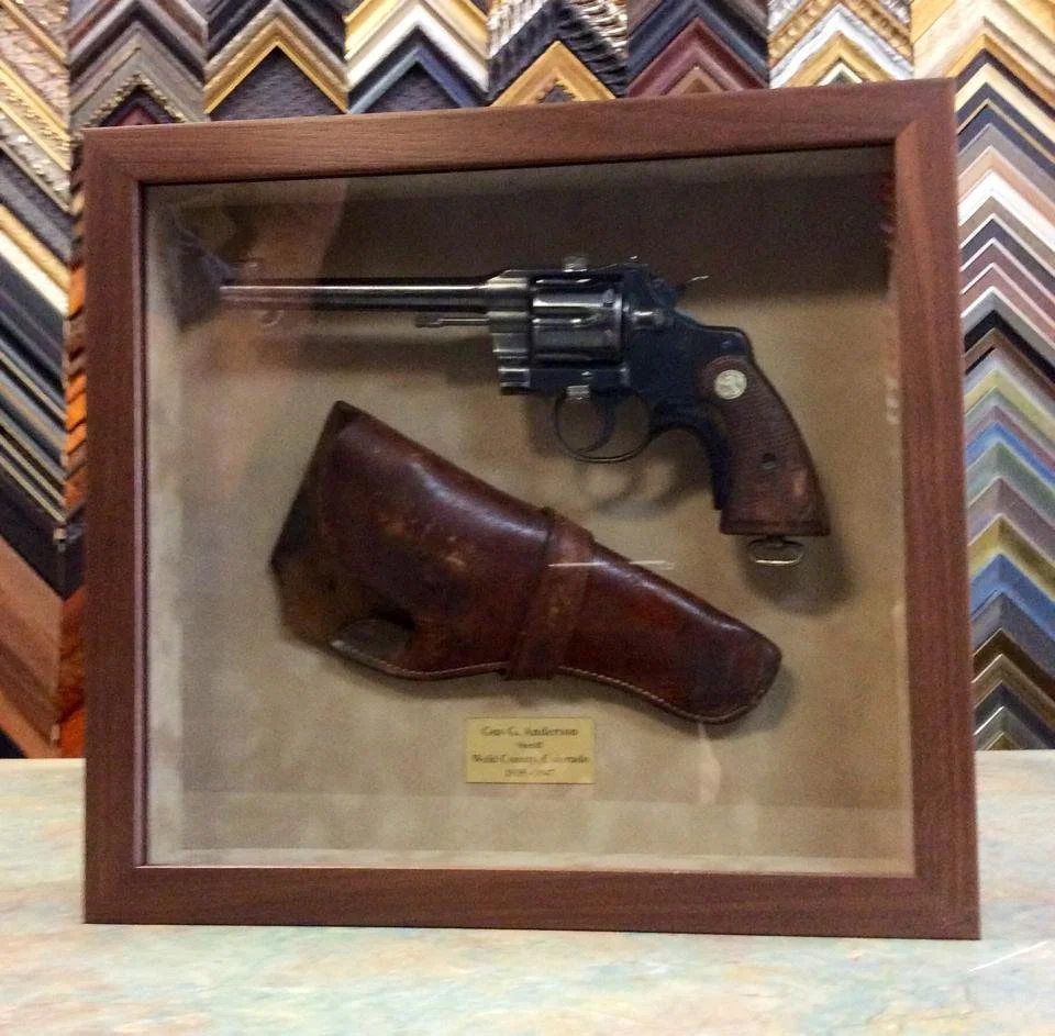 A framed display of a vintage revolver with a brown leather holster underneath, against a background of various picture frame samples.