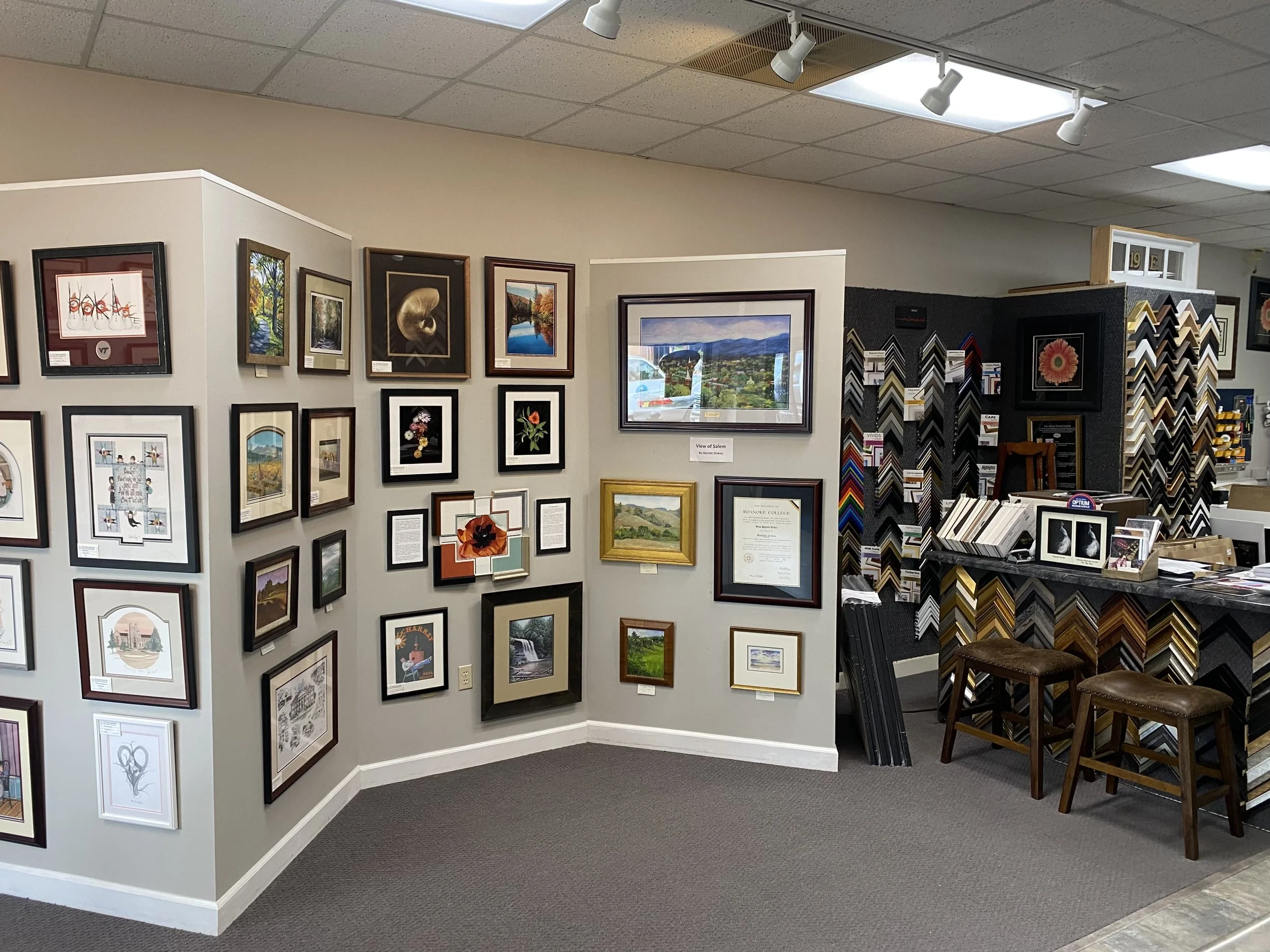 Art gallery with framed paintings and photographs on white and black walls, display counter with framed artworks, and two wooden stools, interior with gray carpet and ceiling lights.