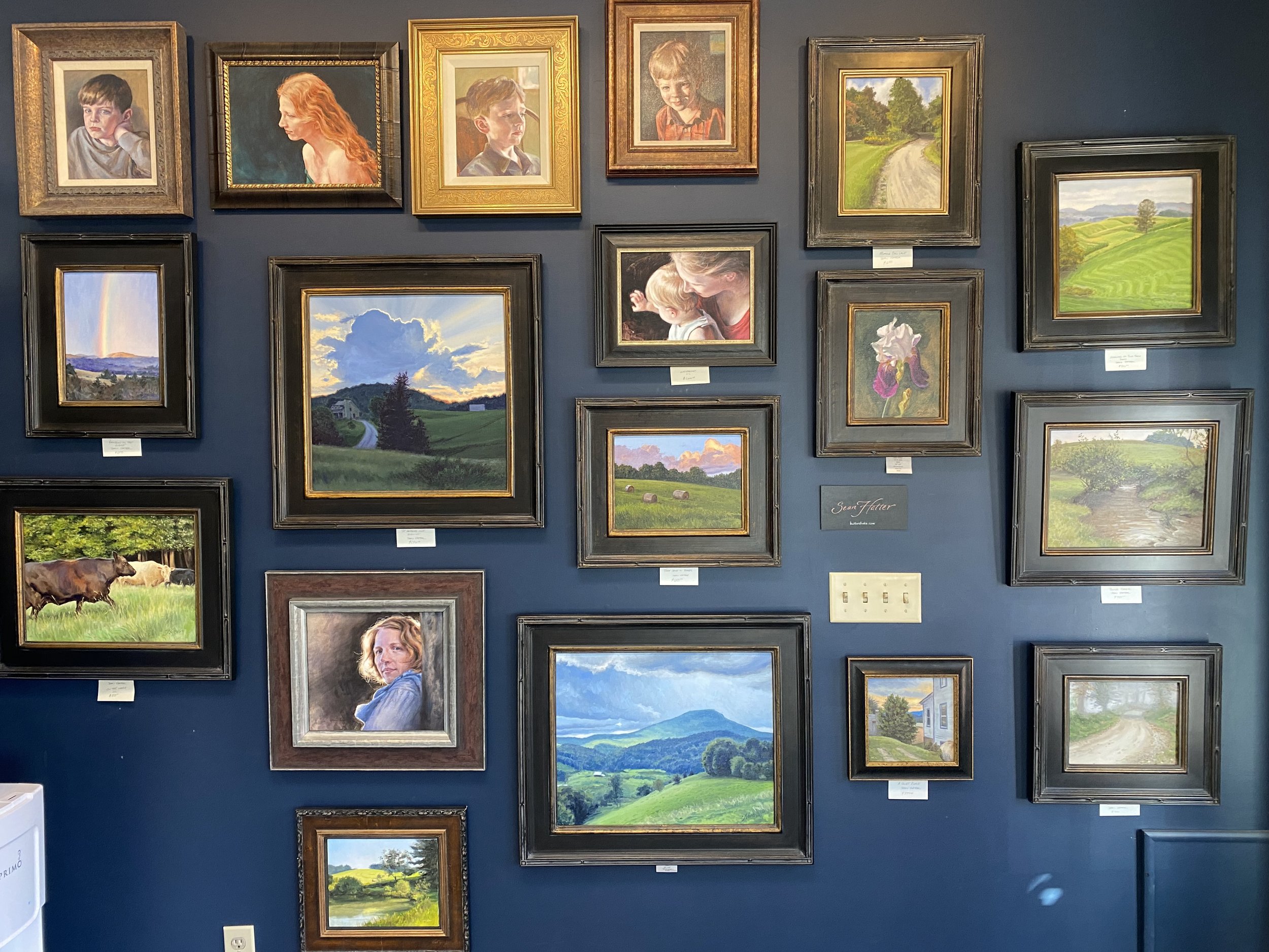 Blue gallery wall displaying various paintings of landscapes, flowers, and portraits.