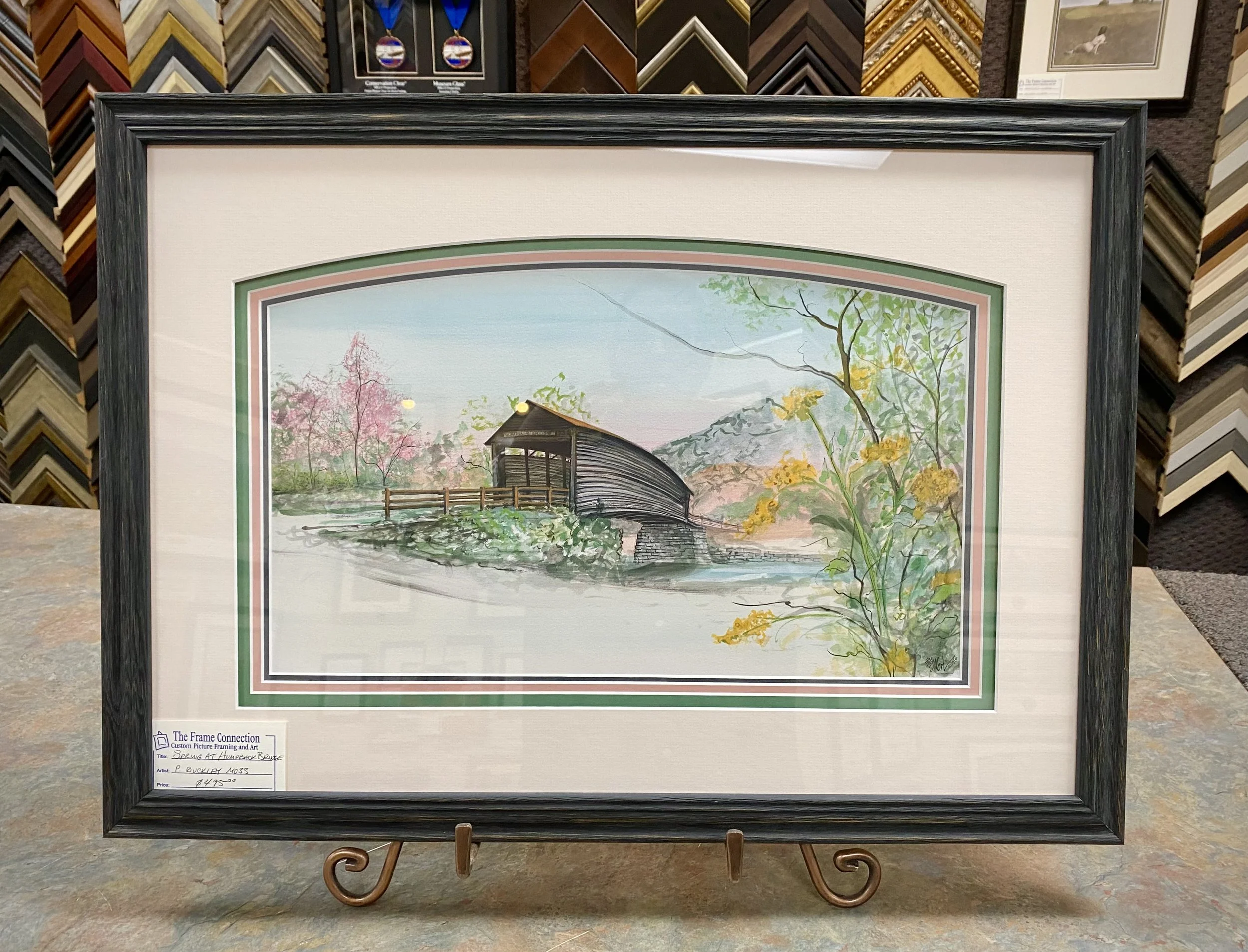 Framed watercolor landscape painting of a covered bridge over a stream, with trees and mountains in the background, in pastel colors.