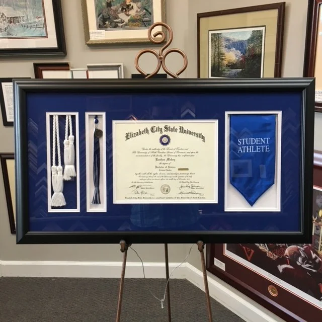 Framed display of an Elizabeth City State University diploma, honor cords and a student athlete pennant on a stand in an art gallery.
