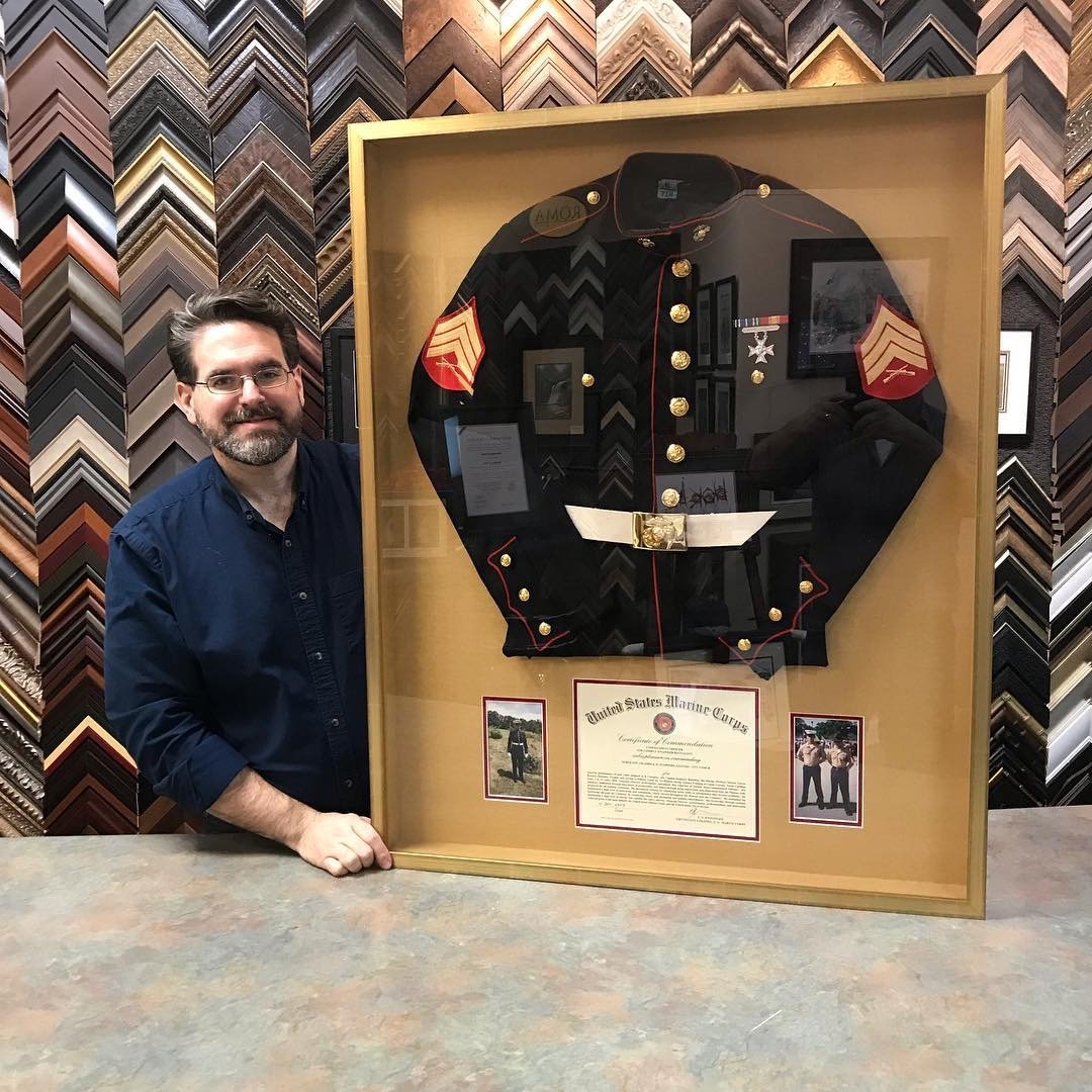 A man with glasses and a beard, wearing a navy blue shirt, stands next to a framed Military Uniform displayed in a glass case. The uniform is a black Marine Corps dress jacket with gold buttons, a red and gold chevron patch on the sleeve, and medals.