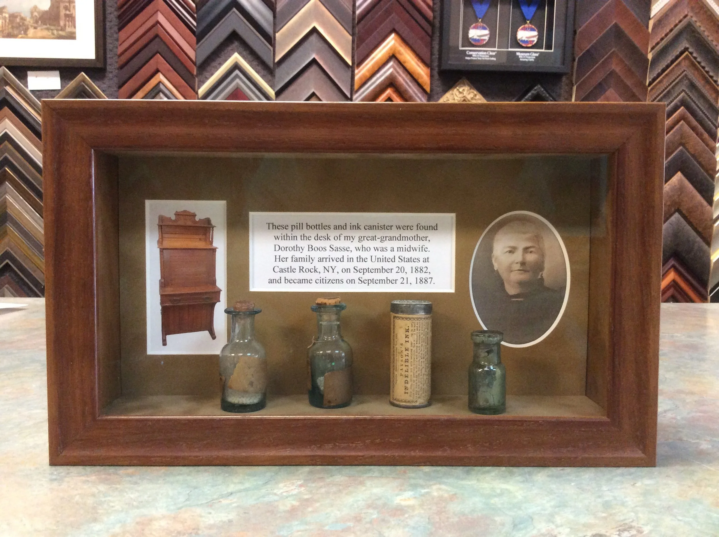 Display case holding vintage bottles and a can, with informational plaque about Dorothy Boos Sasse and her family, and framed artwork and framed medal in the background.
