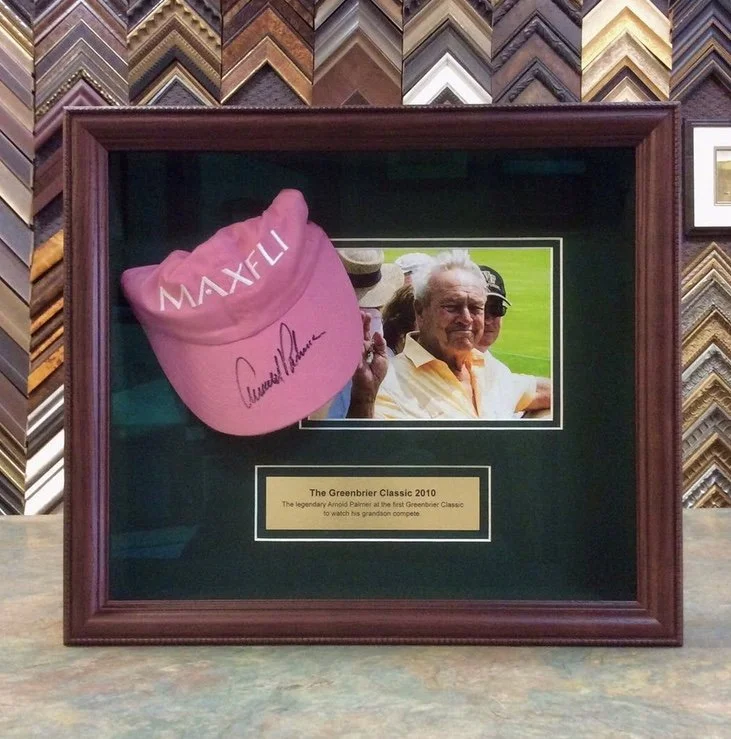 A framed memorabilia display from The Greenbrier Classic 2010 featuring a signed pink cap, a photo of an elderly man holding a golf club, and a plaque, all set against a backdrop of decorative striped framing.