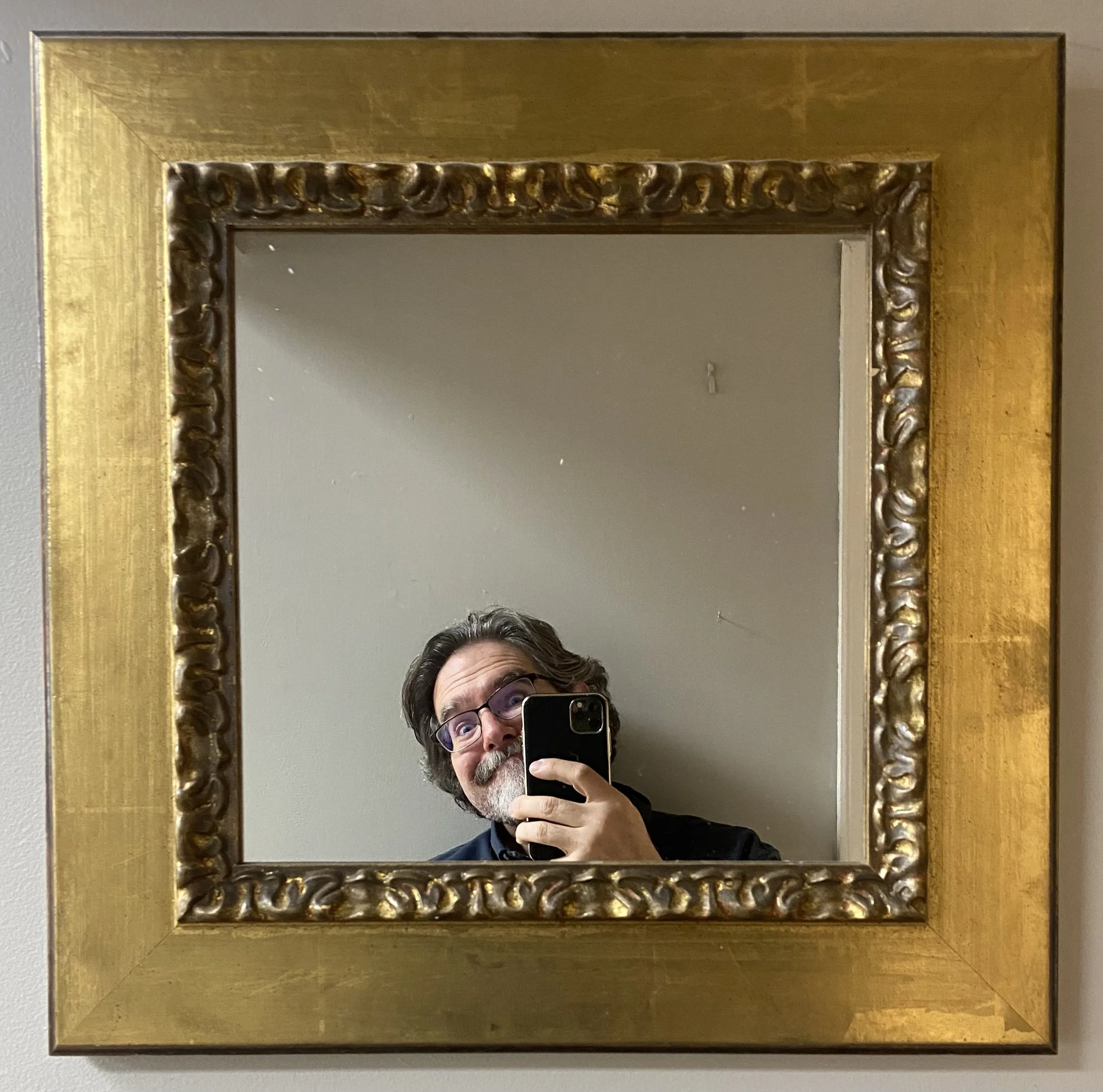 Person taking a mirror selfie, visible face and upper body, in front of a large, ornate gold-framed mirror on a wall. Expert custom mirror framing in Salem, VA, and near Roanoke, VA.